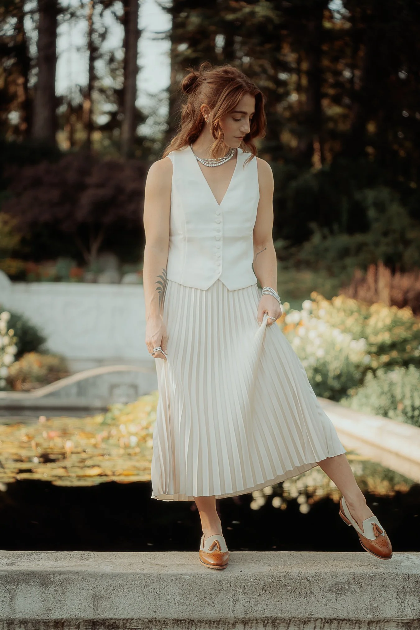 A woman with reddish-brown hair styled in loose waves, wearing a white sleeveless vest, a pleated cream-colored midi skirt, white and brown shoes, and pearl jewelry, standing outdoors near a pond with trees in the background.