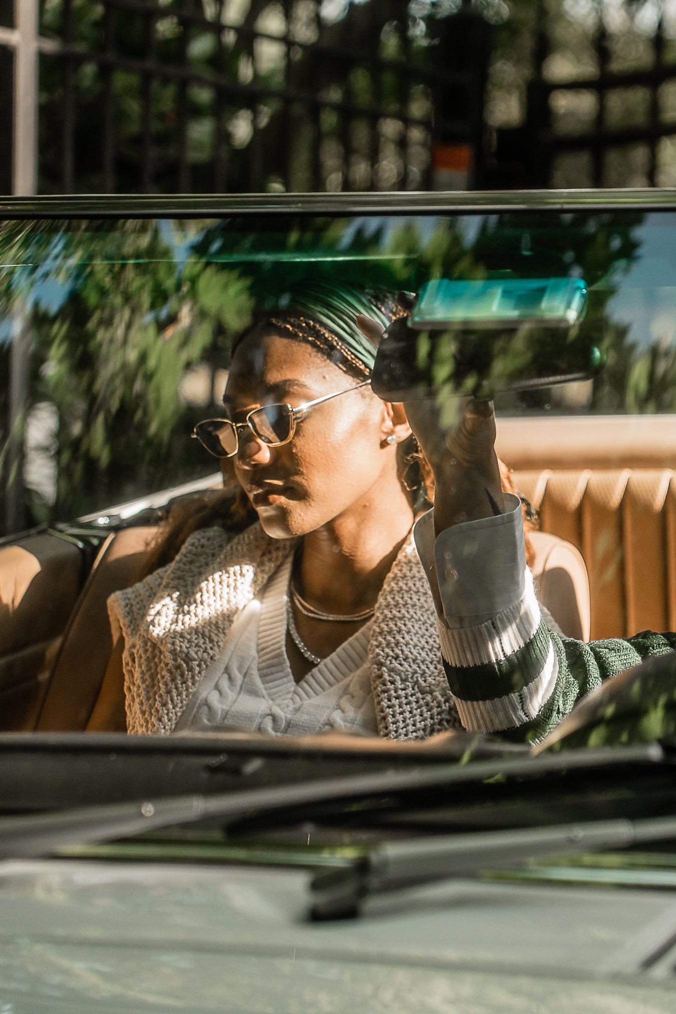 A woman with braided hair, sunglasses, and jewelry sits inside a car, taking a photo of herself through the windshield with her phone. The sunlight highlights her face and outfit.