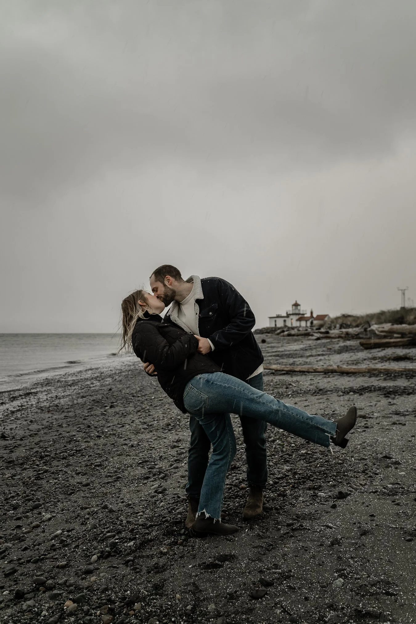 A couple kissing on a rocky beach in front of a lighthouse on a cloudy, rainy day. The man is holding the woman and leaning her back, she has one leg lifted and they are both dressed in dark, casual clothing.