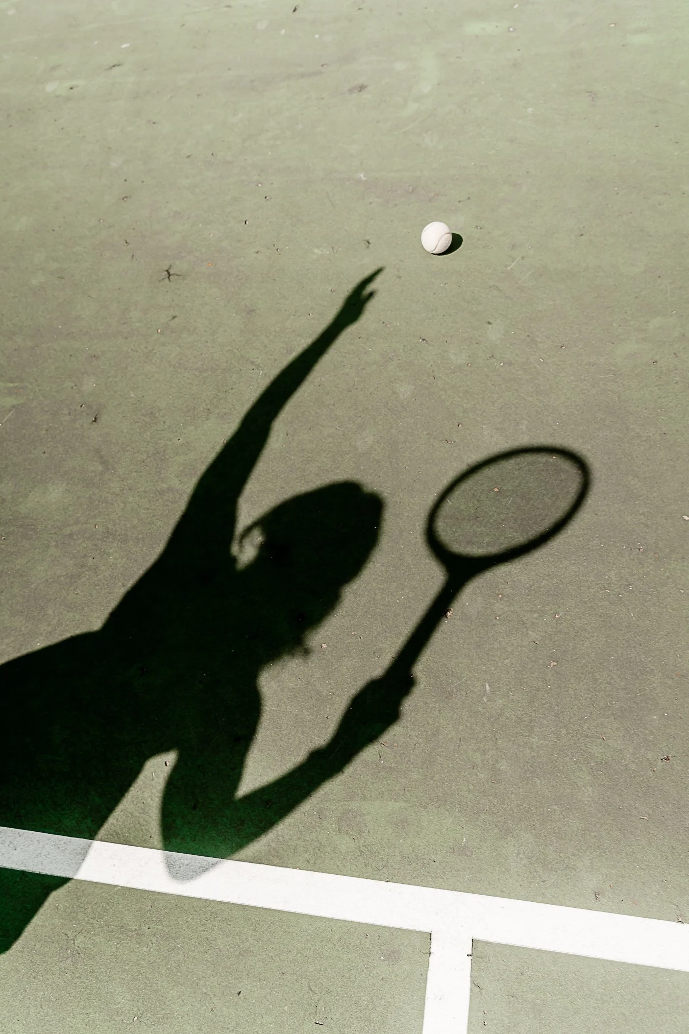 Shadows of a person reaching for a tennis ball and holding a tennis racket cast on a tennis court.