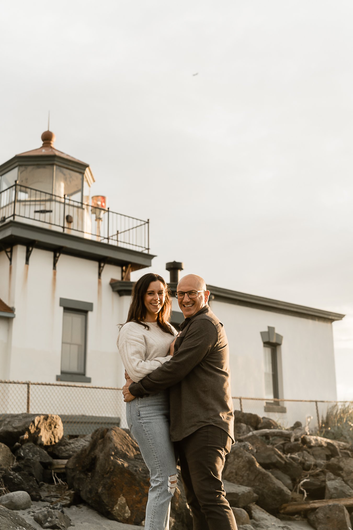 A smiling couple stands together outdoors near a rocky shoreline with a white lighthouse building in the background during sunset.