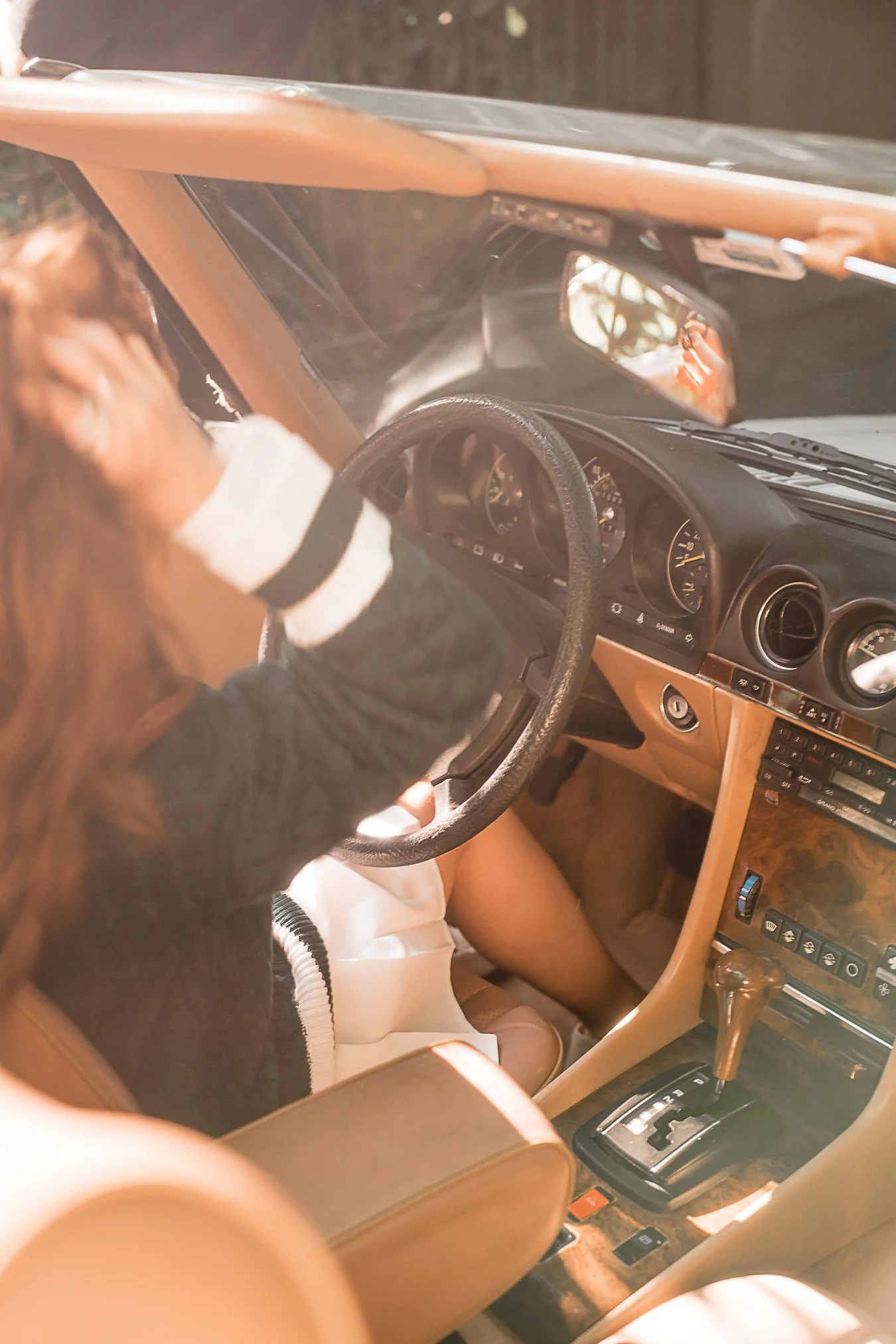 Interior of a vintage car with a woman wearing a black sweater and white skirt, holding the steering wheel. The dashboard and gear shift are visible with wood paneling accents.