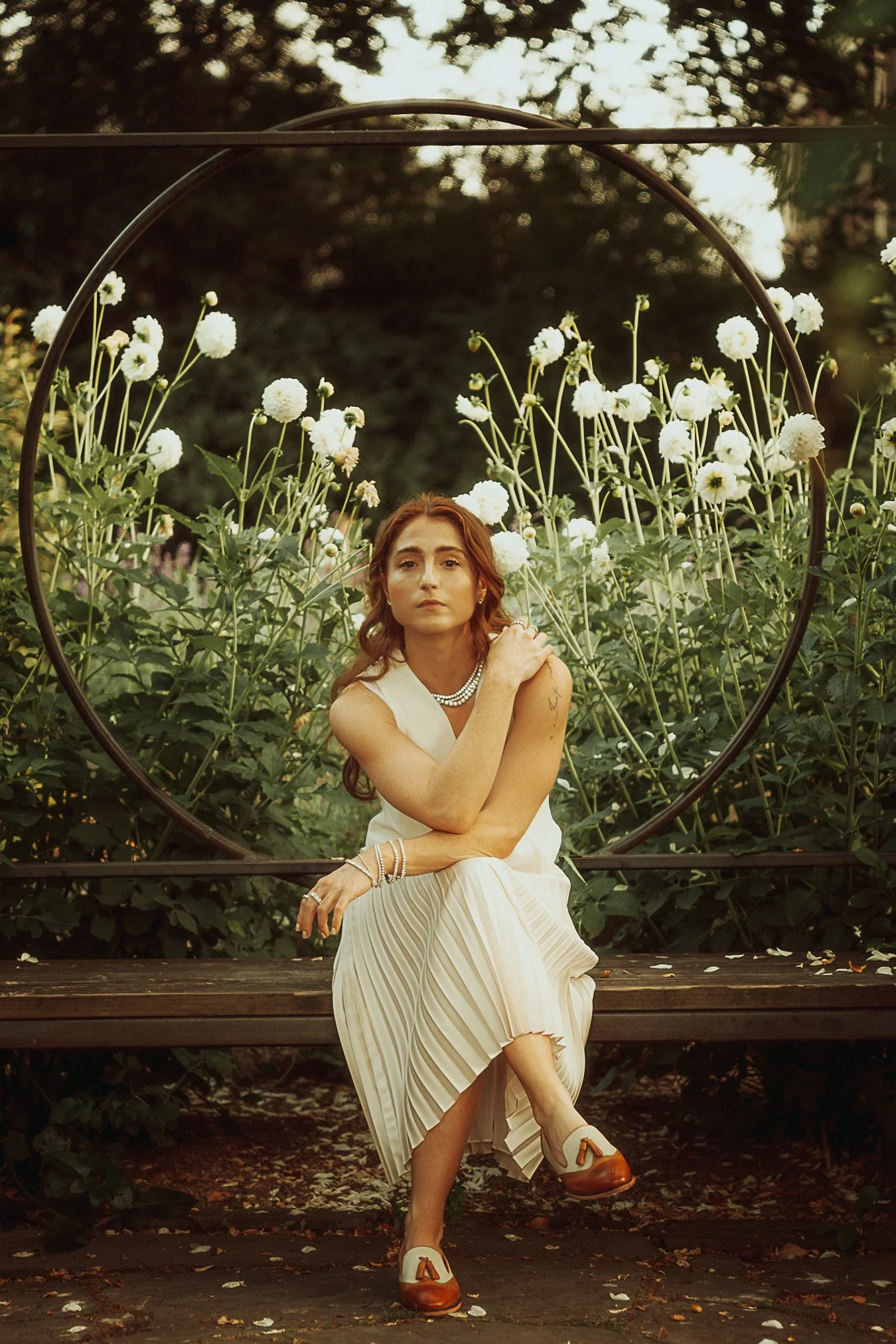 A woman with red hair, dressed in a white pleated skirt and top, sitting on a park bench surrounded by white flowers and greenery at sunset, looking thoughtfully at the camera.