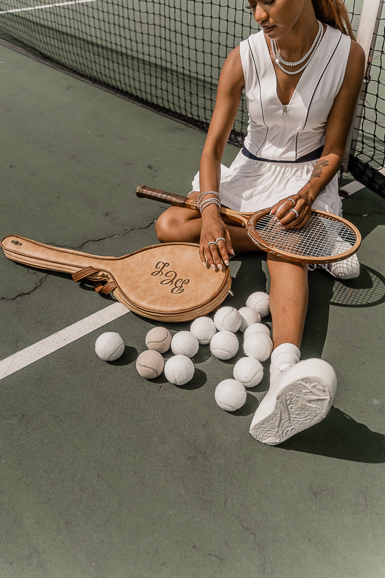 A woman in tennis attire sitting on a tennis court with tennis balls, a tennis racket, and a guitar case around her.