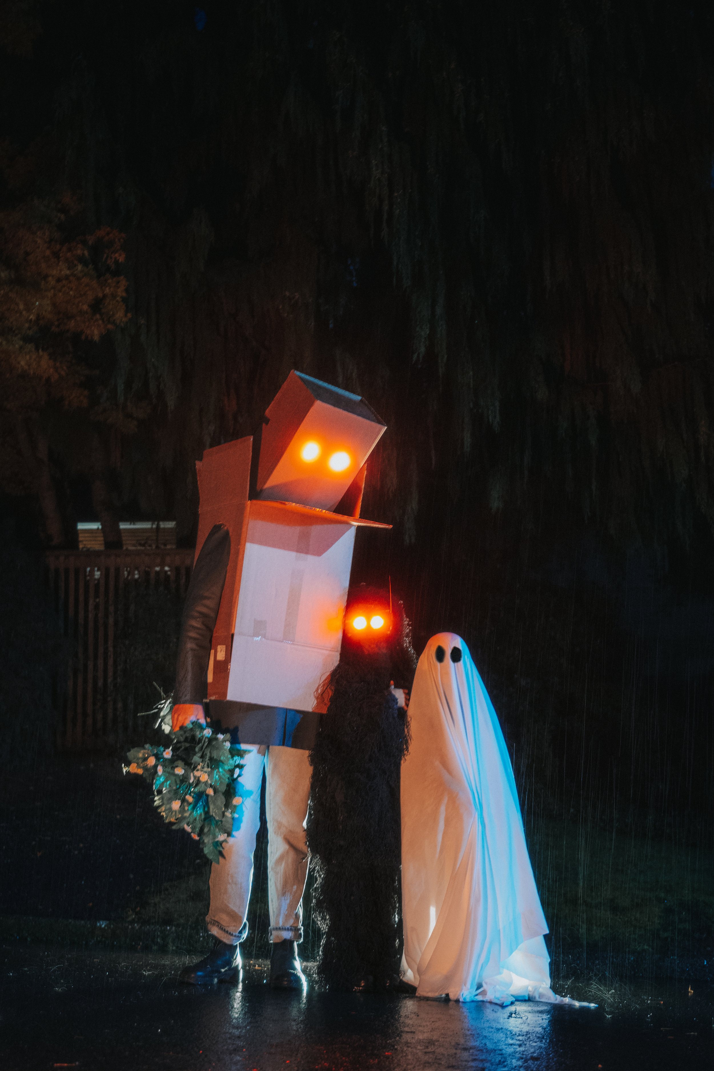 People in costumes standing outside at night, with a large tree in the background. One person is dressed as a pumpkin, another as a ghost, and another in a dark furry costume. The pumpkin costume has glowing eyes and the ghost costume has a white she