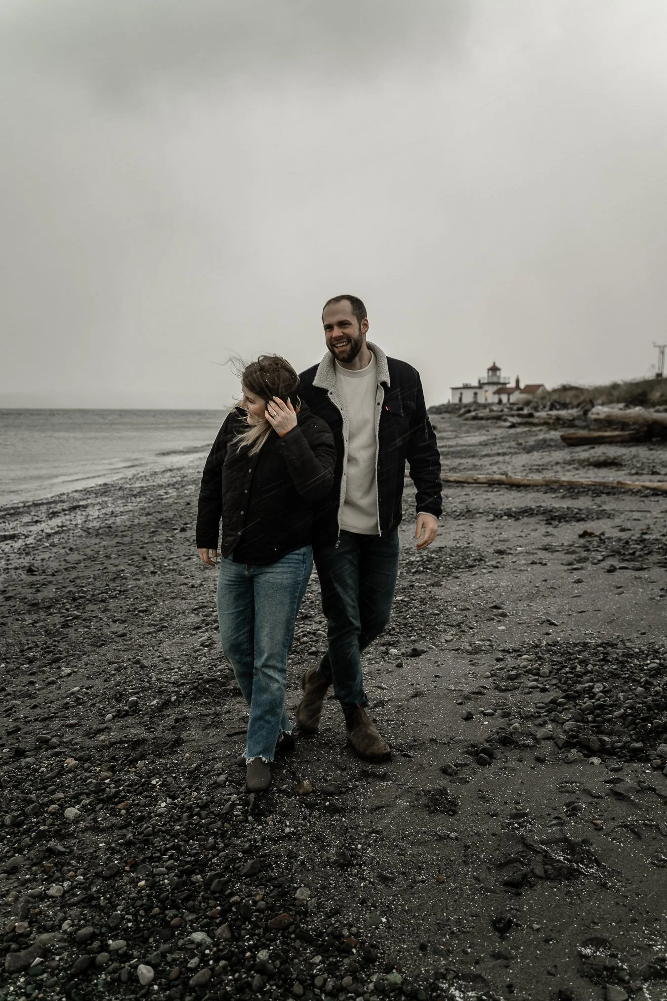 A couple walking on a rocky beach with a lighthouse in the distance, overcast sky, and wearing dark jackets.
