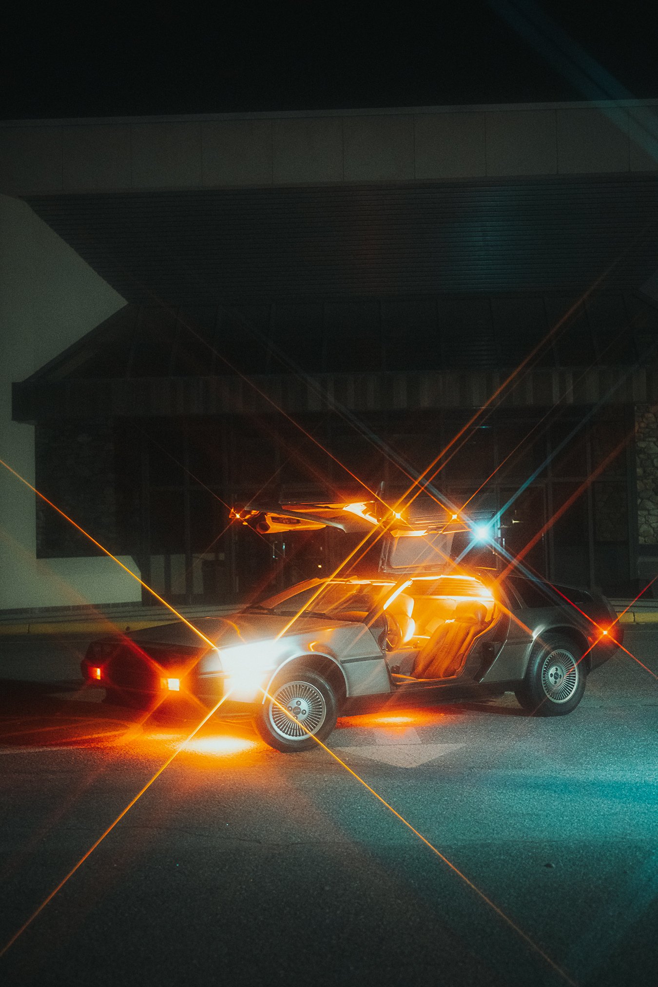 A DeLorean sports car parked in front of a modern building at night, illuminated by bright lights creating starburst effects.