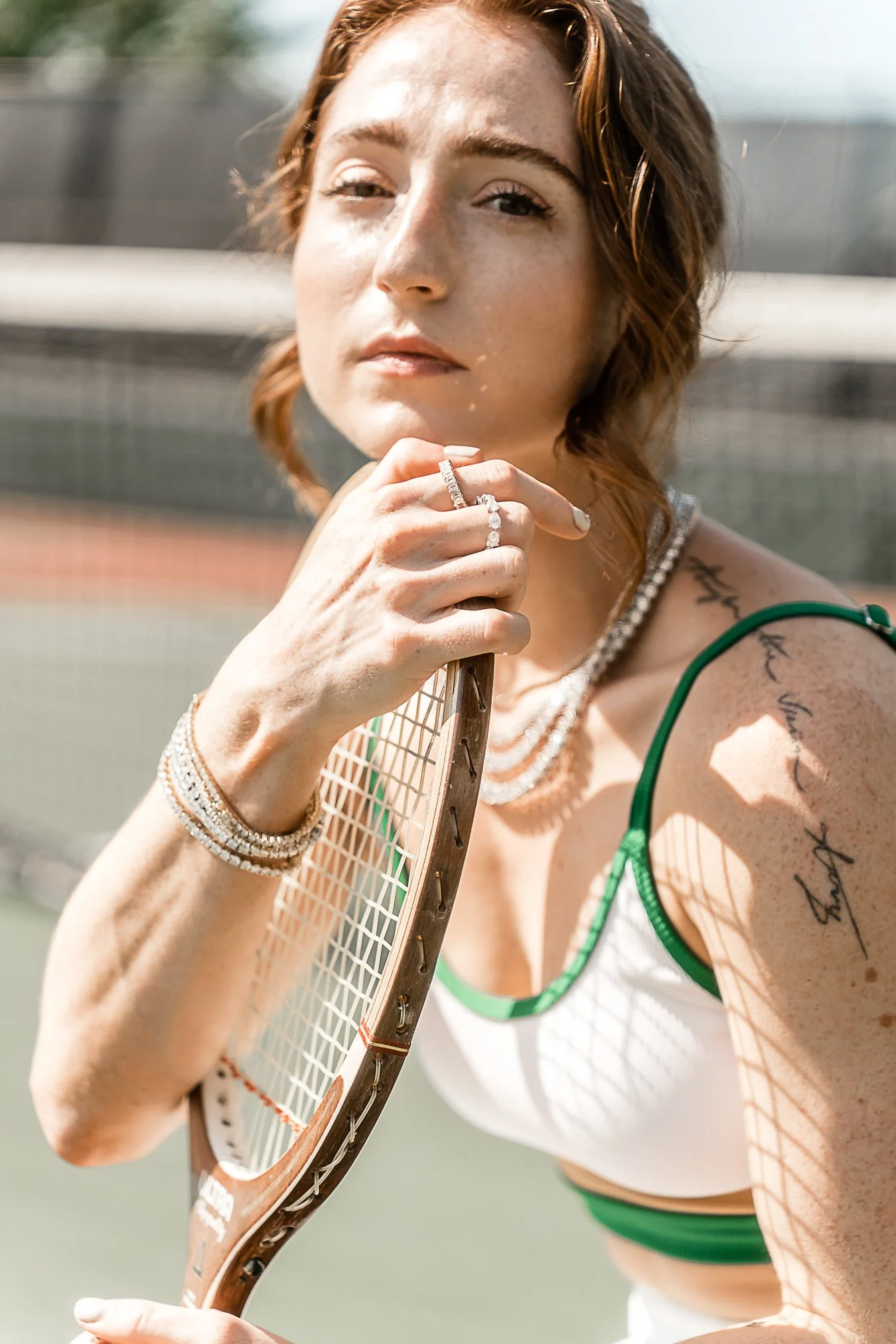 A woman with freckles holding a tennis racket near her face on a tennis court, wearing jewelry and a white and green sports outfit.