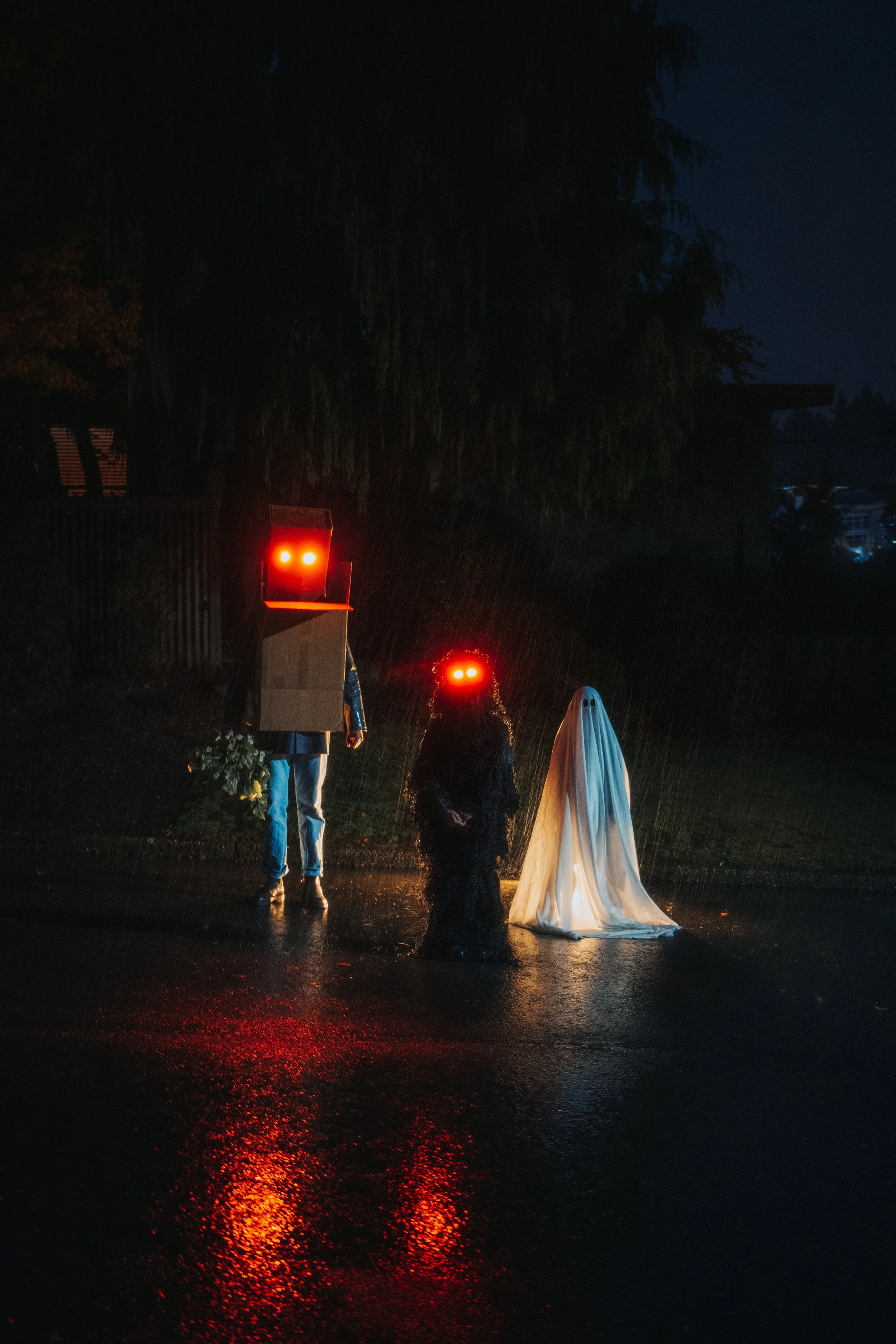 Three people dressed in Halloween costumes standing on a rainy street at night, with red lights reflecting on the wet pavement.