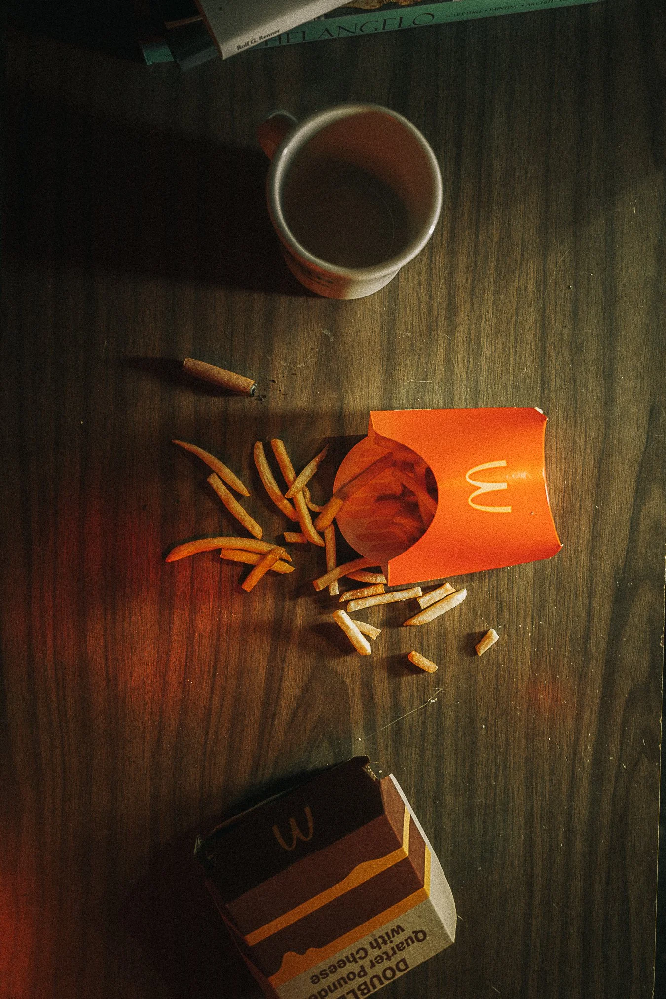 A spilled box of McDonald's french fries, a cup of coffee, and a pack of Dutch Gold cigarettes on a wooden table, with a magazine partially visible in the background.