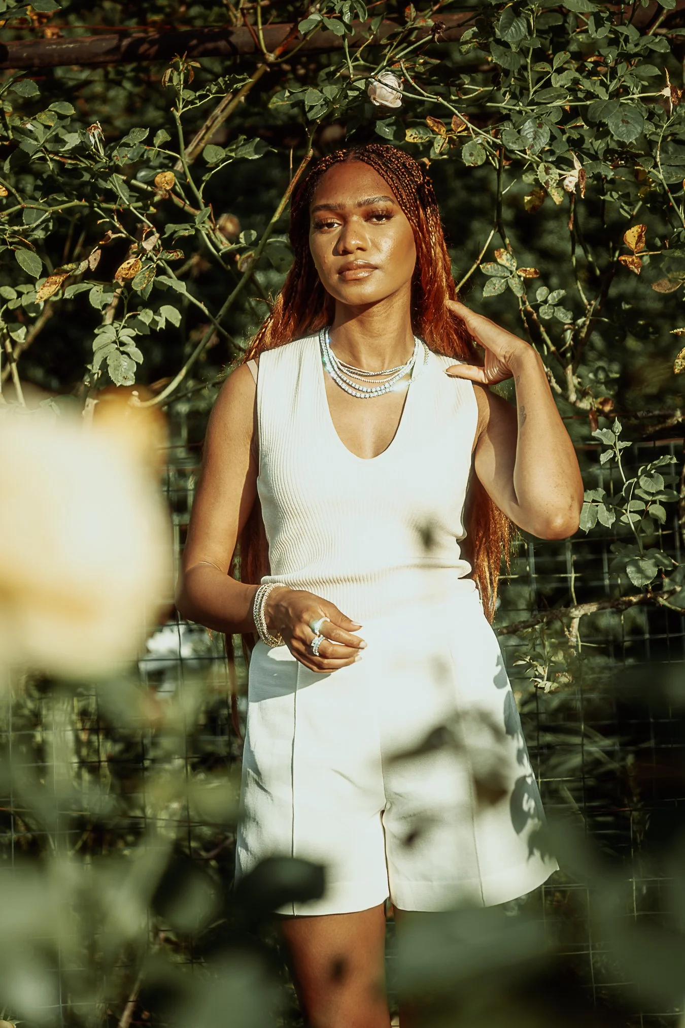 A woman with long braids, dressed in a white sleeveless top and matching white shorts, standing outdoors among green foliage and rose bushes, wearing layered necklaces and bracelets, with the sunlight highlighting her face.