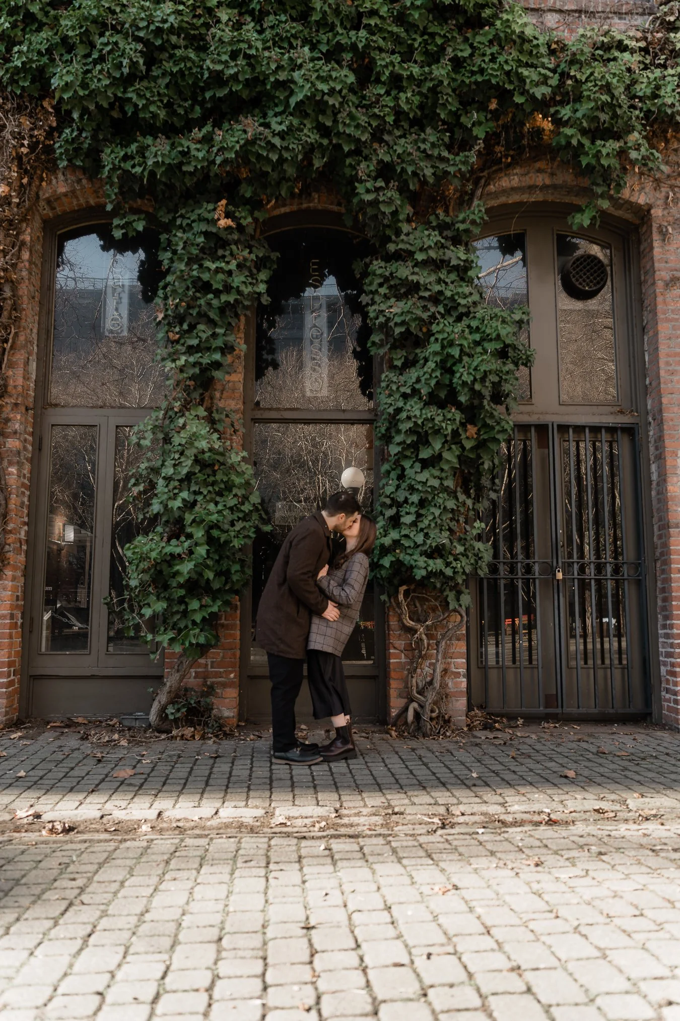 A couple kissing in front of a building covered with green ivy, with brick and glass windows.