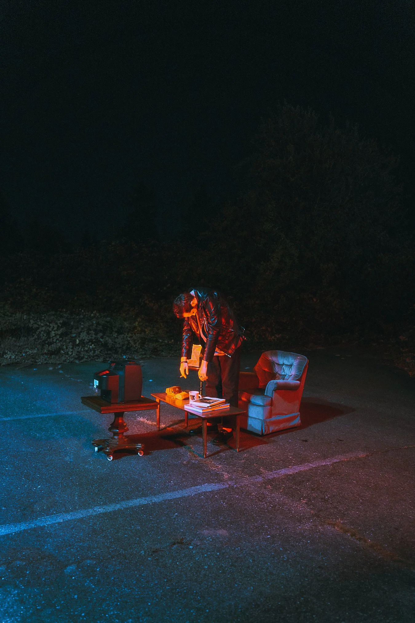 A person in a leather jacket standing in a parking lot at night, illuminated by red and blue lights, with a coffee mug, books, and electronic devices on a table, and an armchair nearby.