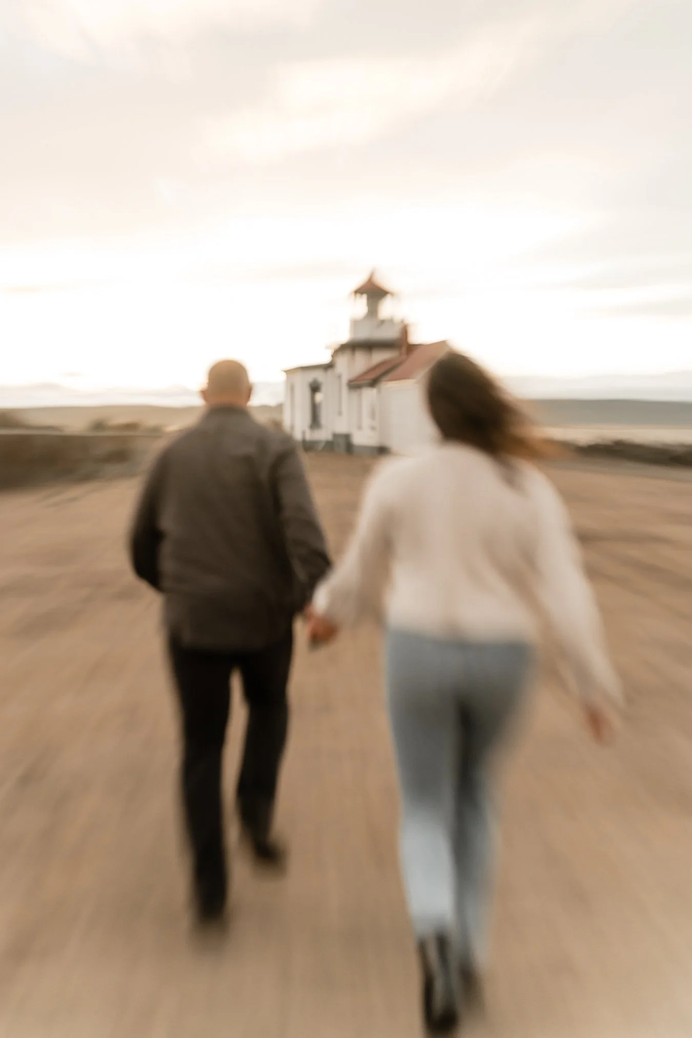 A couple walking hand in hand on a dirt path near a white lighthouse at sunset.