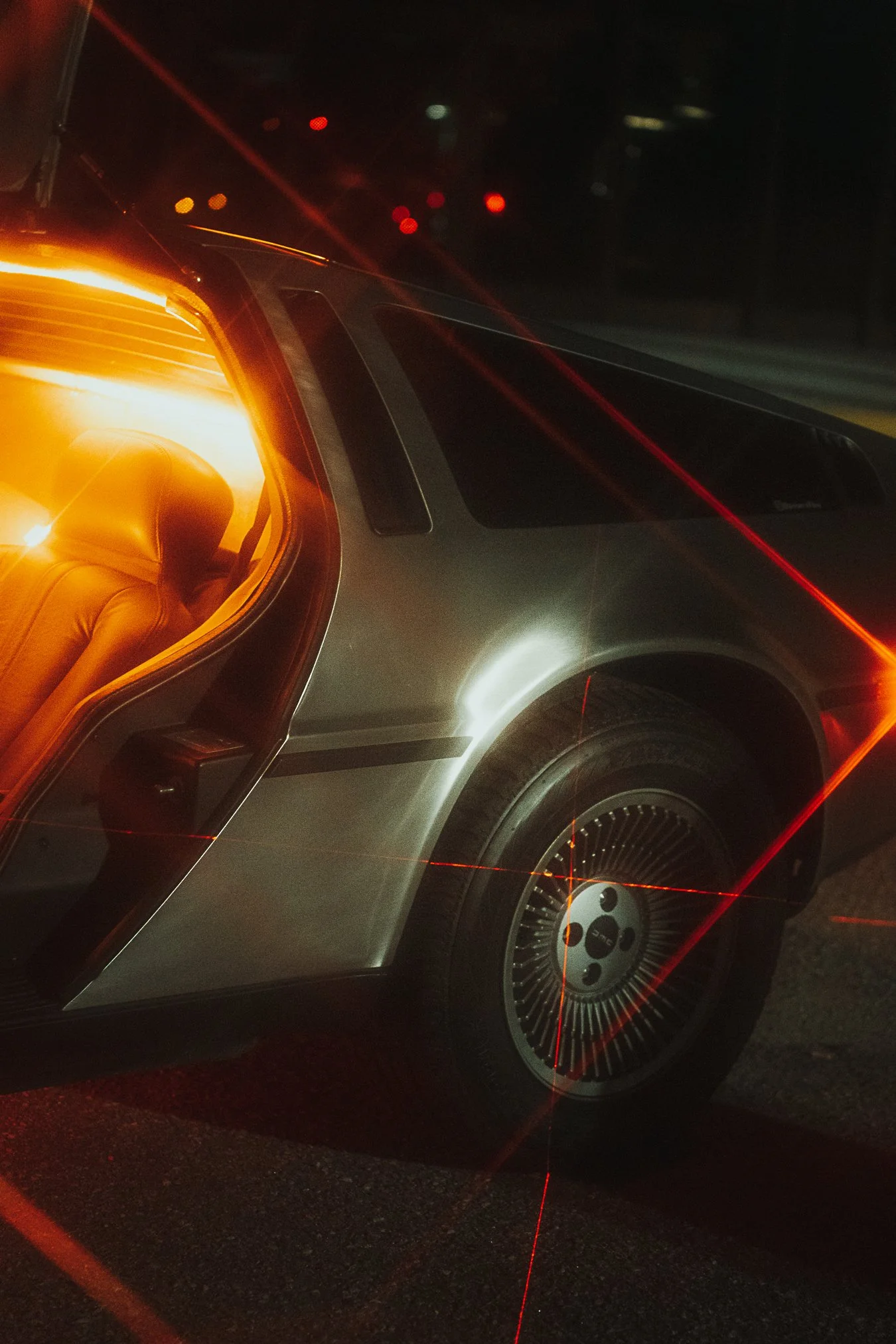 A deLorean car with its gull-wing door open, illuminated by the interior orange/yellow light, has laser-like red lines crossing over the image, set against a dark background.