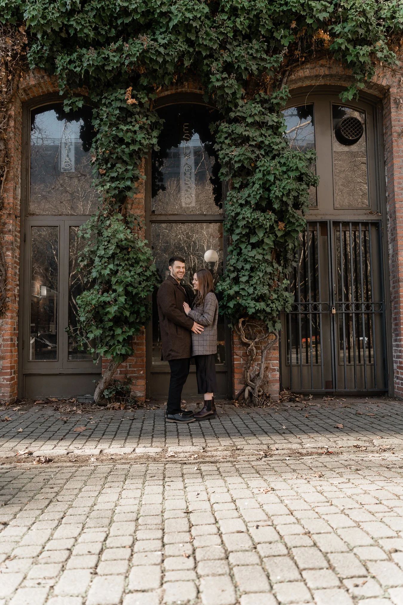 A man and woman standing close together on a cobblestone sidewalk outside a building with large windows and ivy-covered brick walls, smiling at each other.