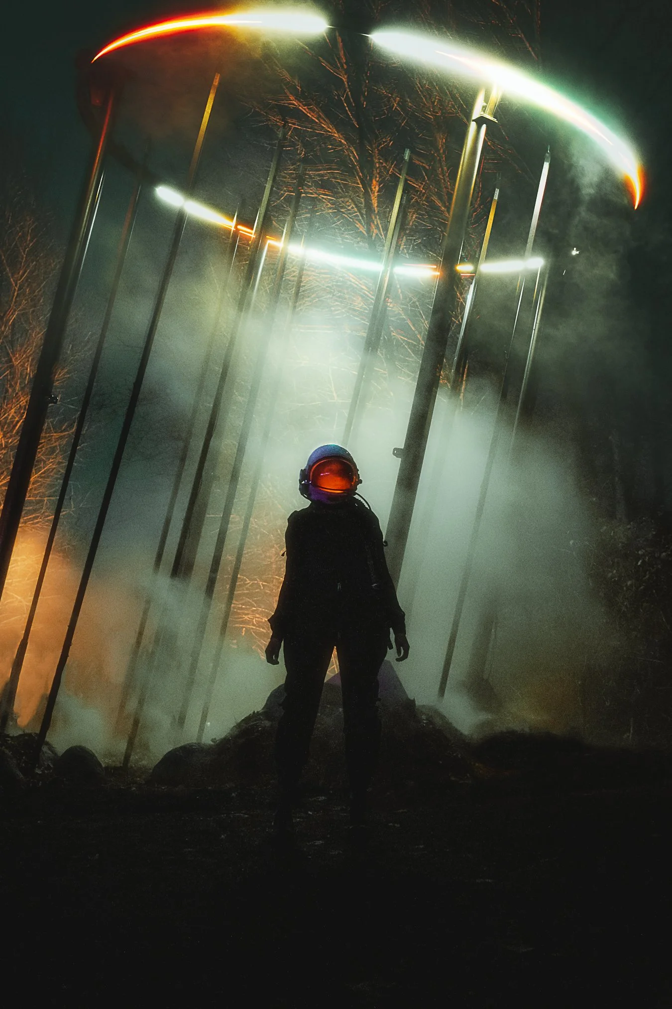 A person wearing a helmet and dark clothing stands on a trail at night, surrounded by fog, with tall light poles and smoke in the background.