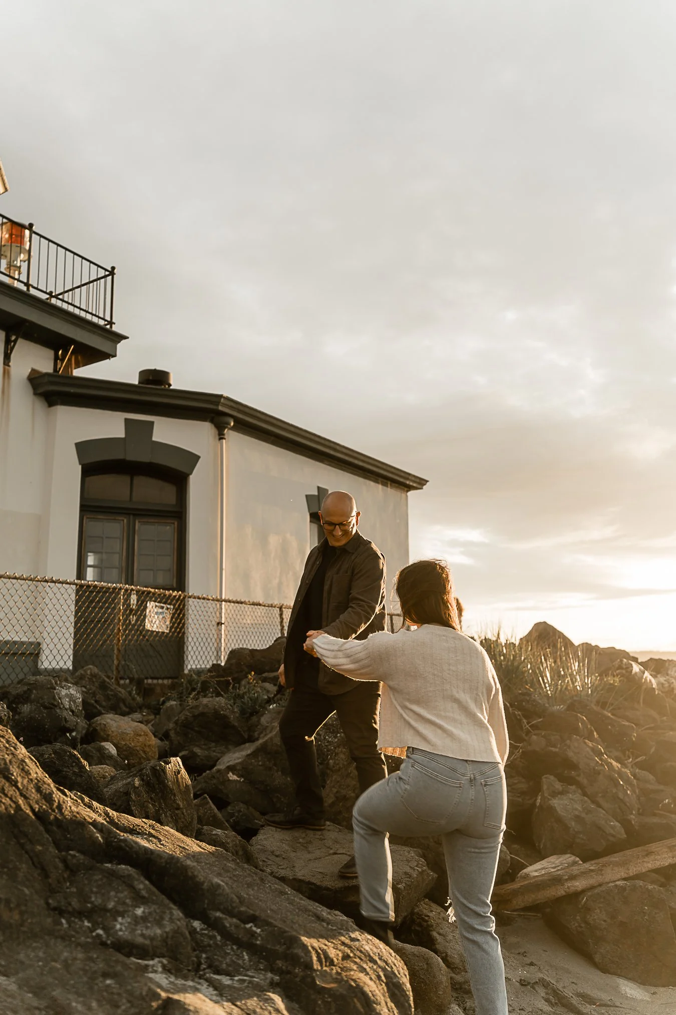 A man is helping a woman up on rocks in front of a house at sunset.