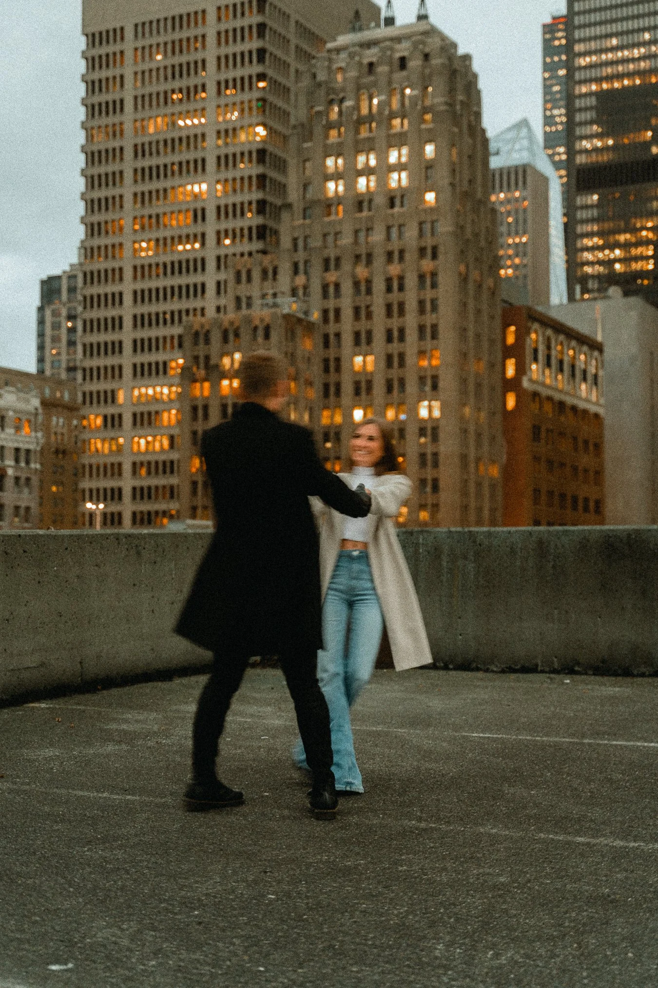 A couple dancing on a rooftop parking lot with city skyscrapers in the background during evening.