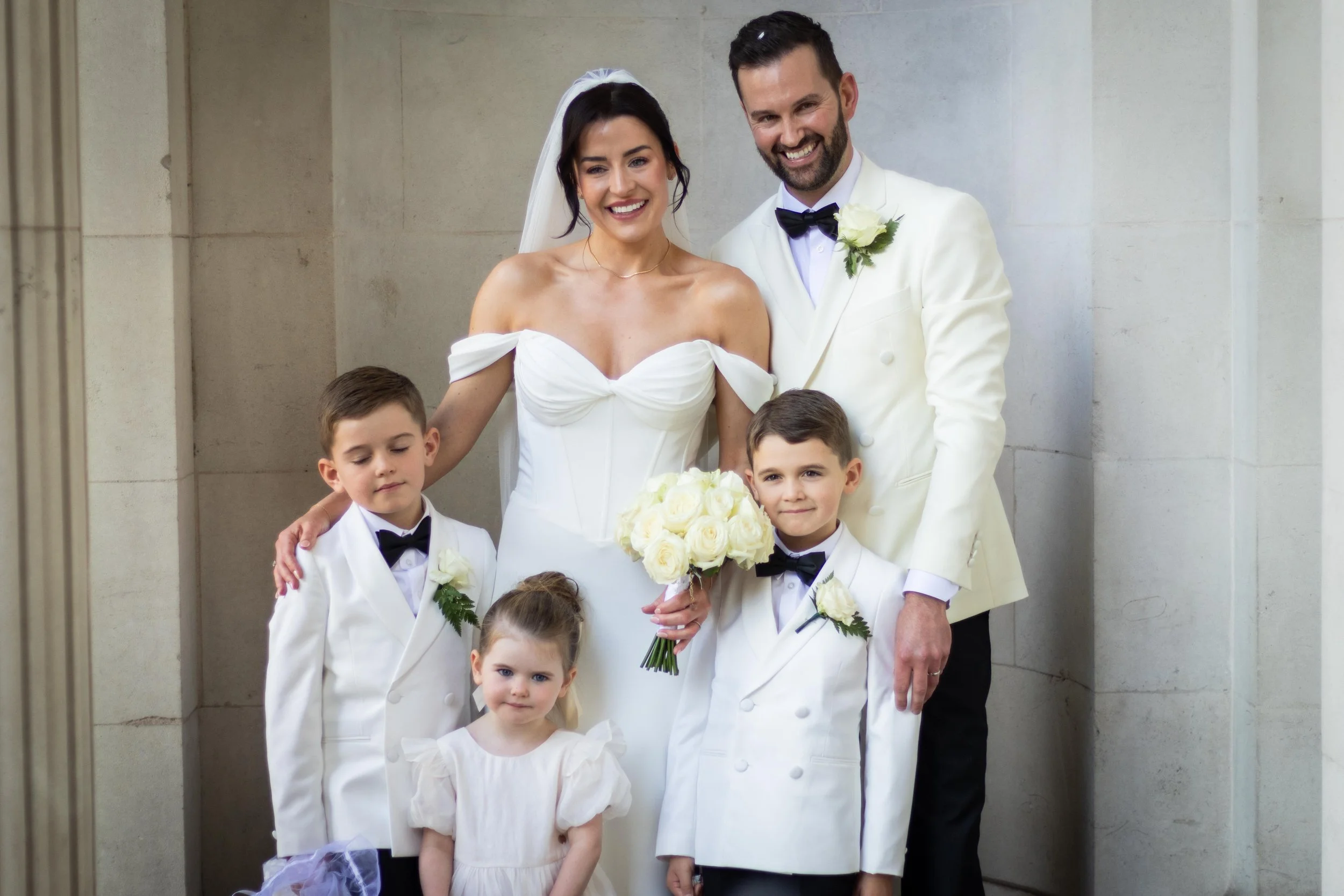 A wedding family portrait featuring a bride and groom with four children, all dressed in formal white and black attire, standing in front of a stone wall.