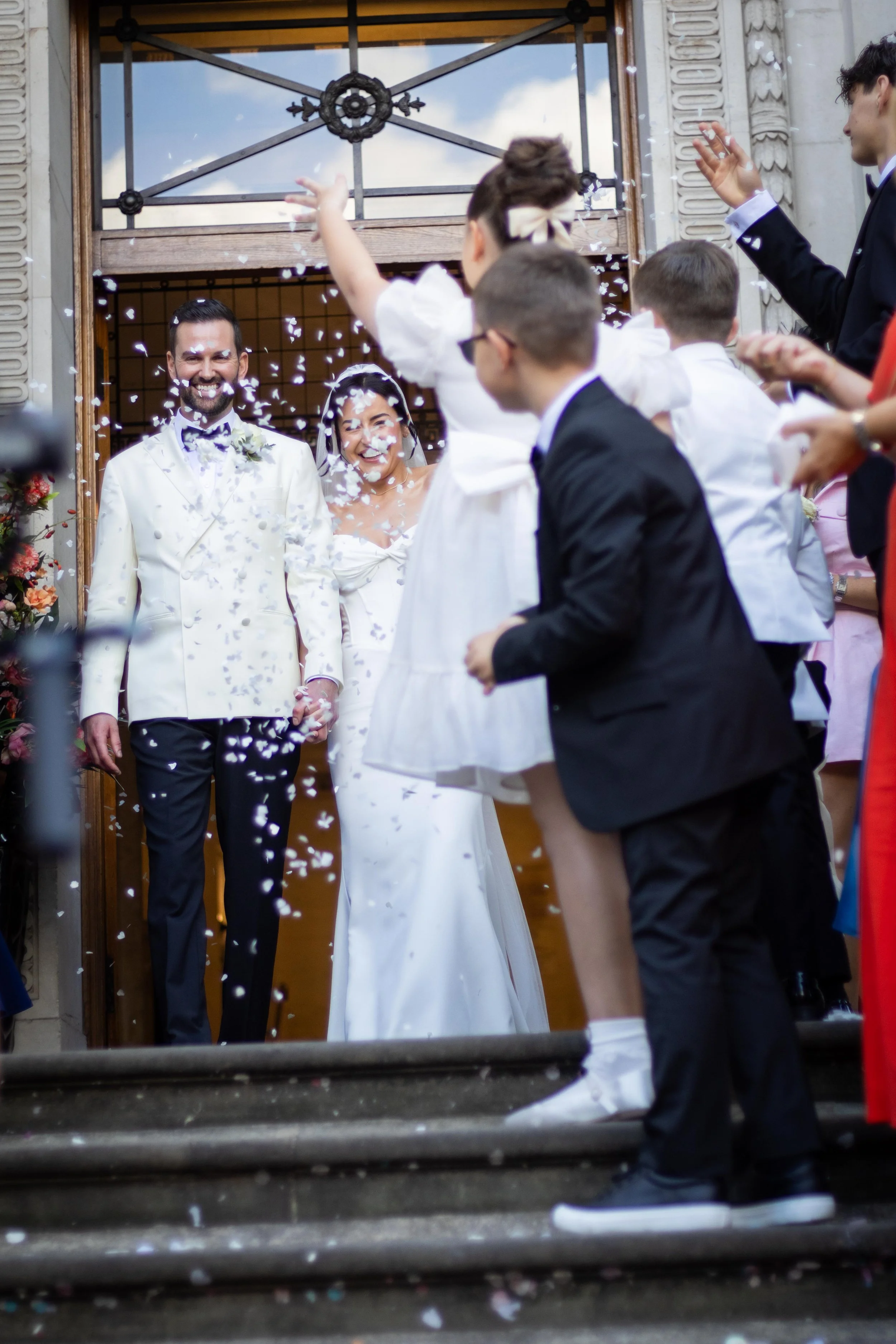 Bride and groom smiling as children and guests celebrate with confetti outside a church during a wedding ceremony.