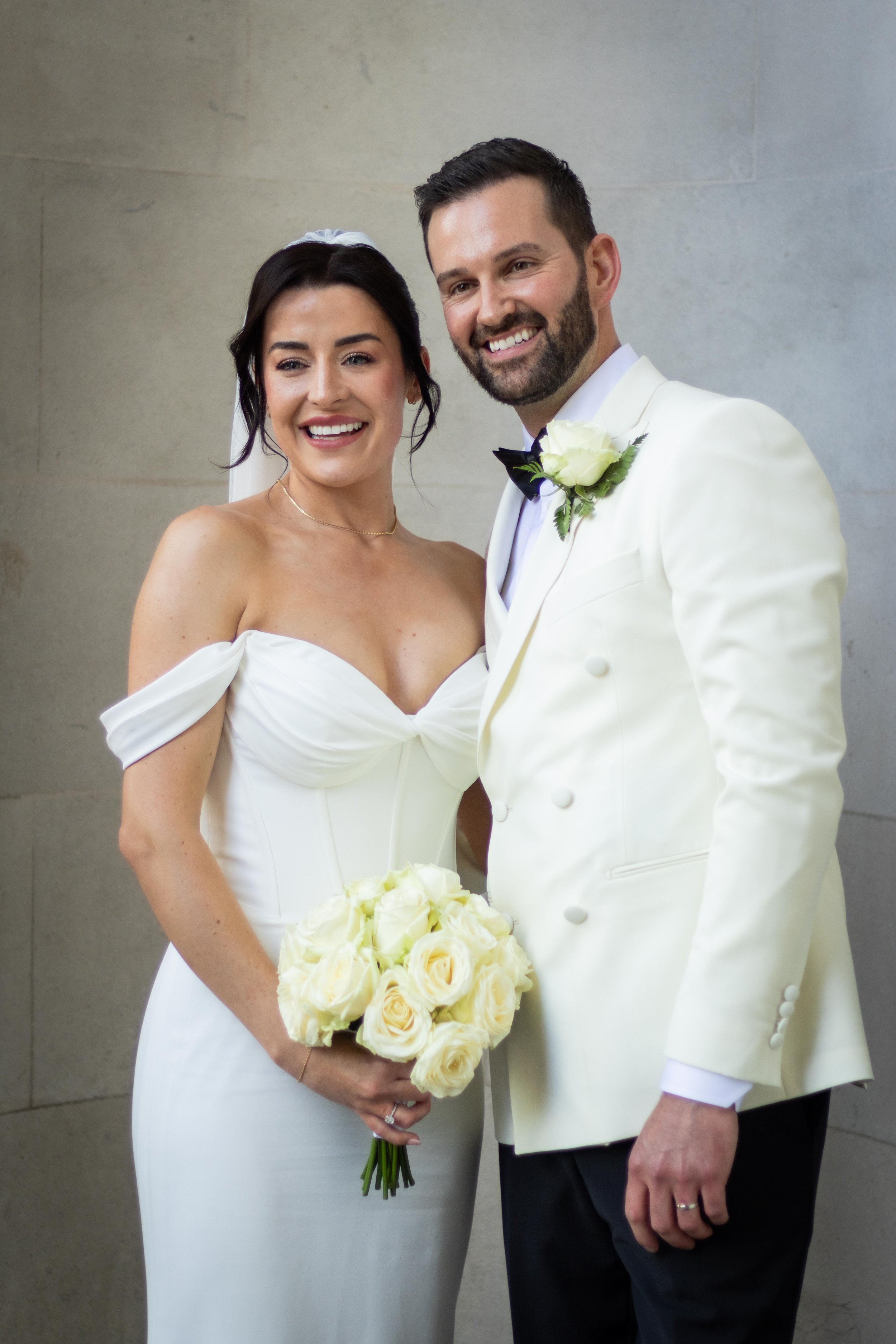 A bride and groom at their wedding, smiling and standing close together. The bride is holding a bouquet of white roses, and the groom is wearing a white tuxedo jacket with a black bow tie and a white rose boutonniere.