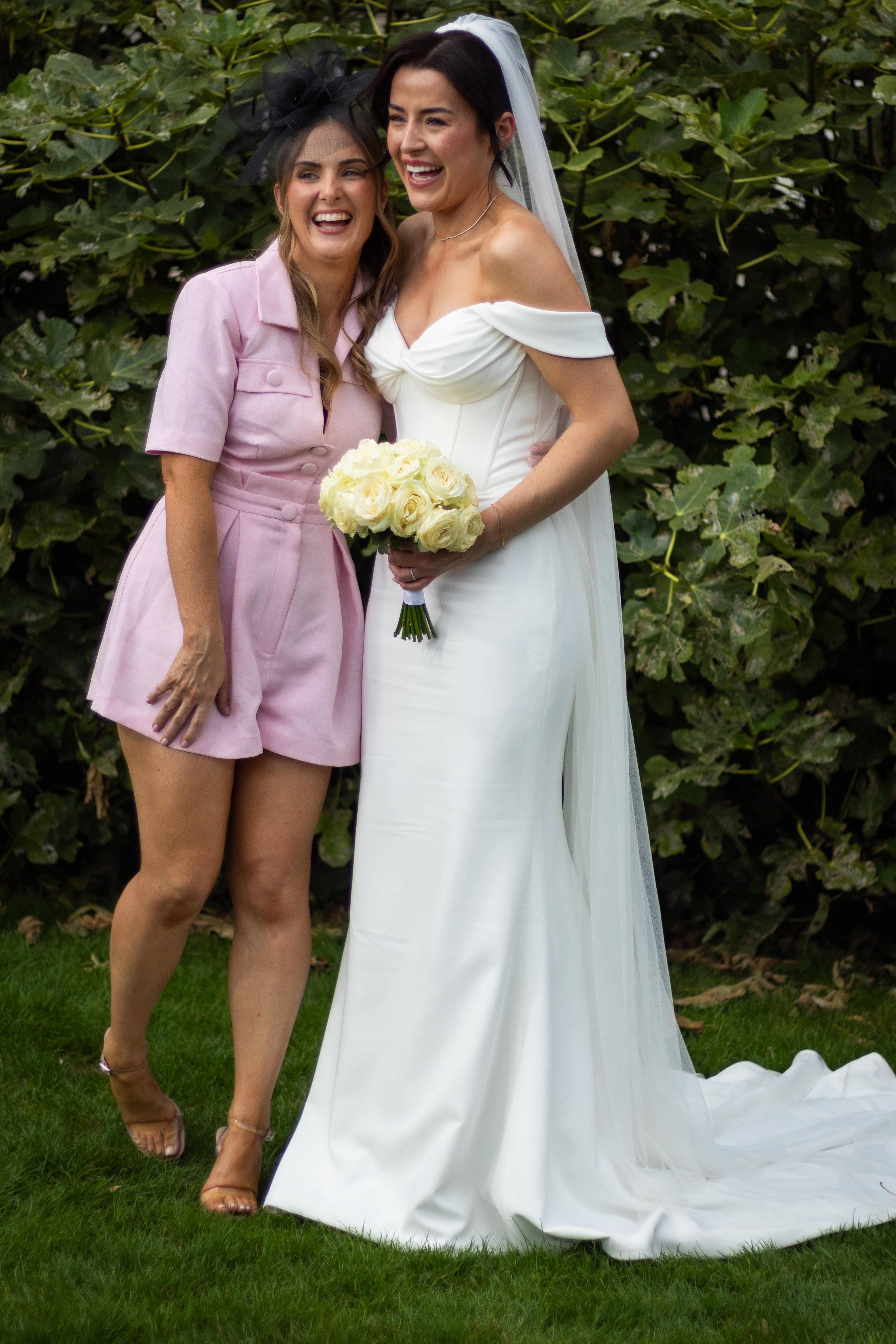 Two women, one in a white wedding dress holding a bouquet of yellow roses, and the other in a pink short suit with a black fascinator, are smiling and hugging outdoors near green bushes.