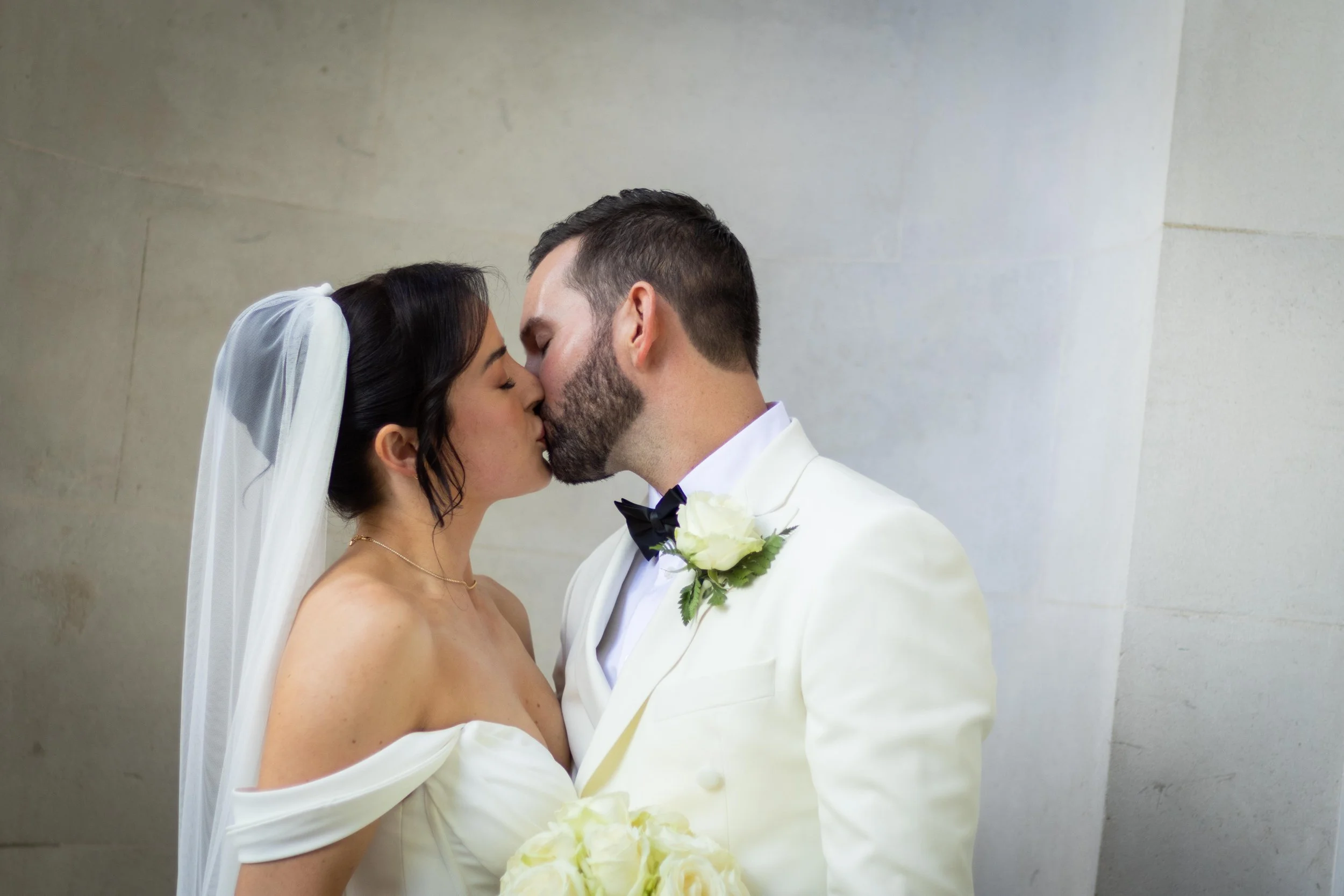 A bride and groom sharing a kiss on their wedding day. The bride is wearing an off-shoulder white wedding dress with a veil, and the groom is in a white tuxedo with a black bow tie and a white flower boutonniere. The bride is holding a bouquet of whi