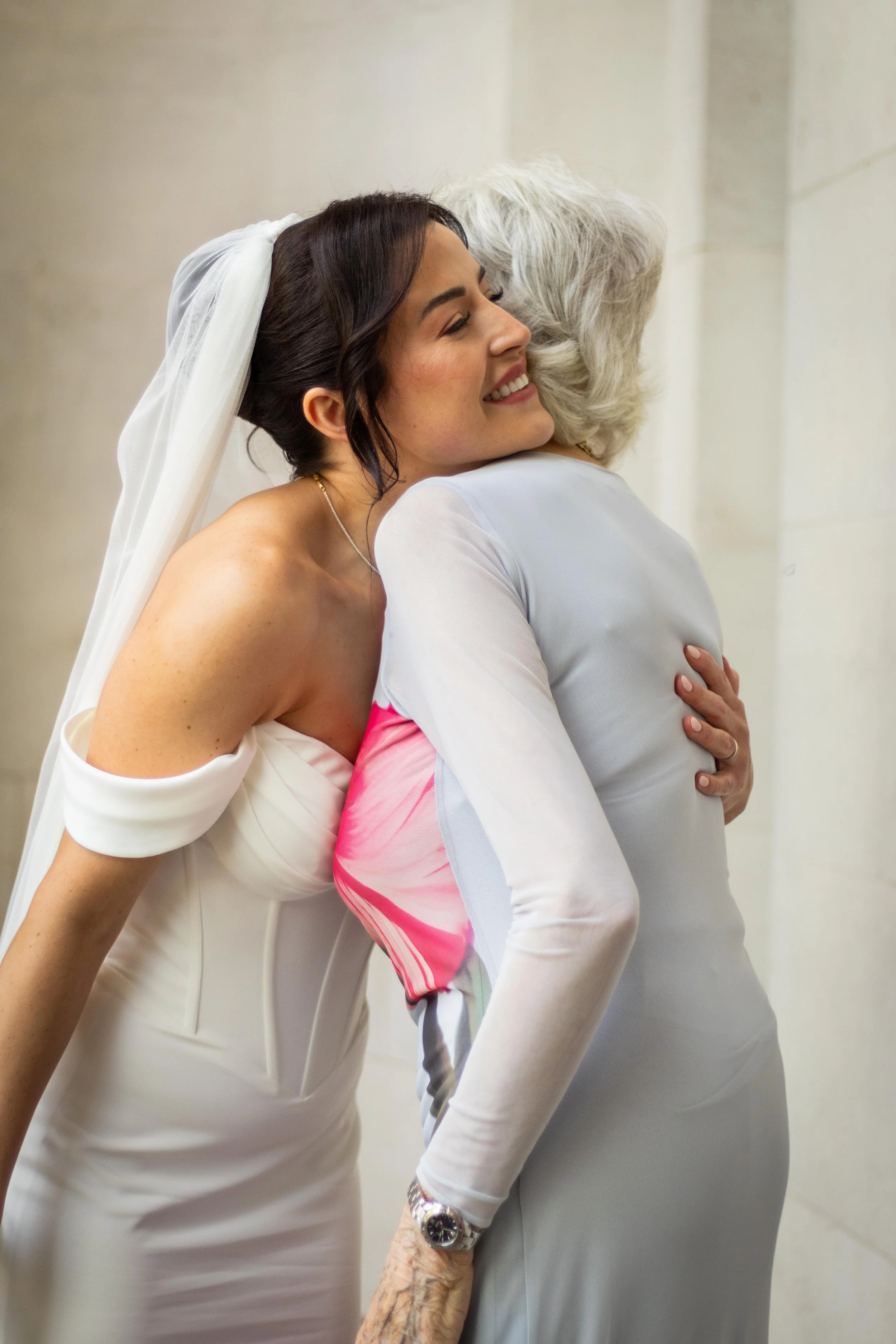 A bride hugging an older woman, both smiling, inside a building.