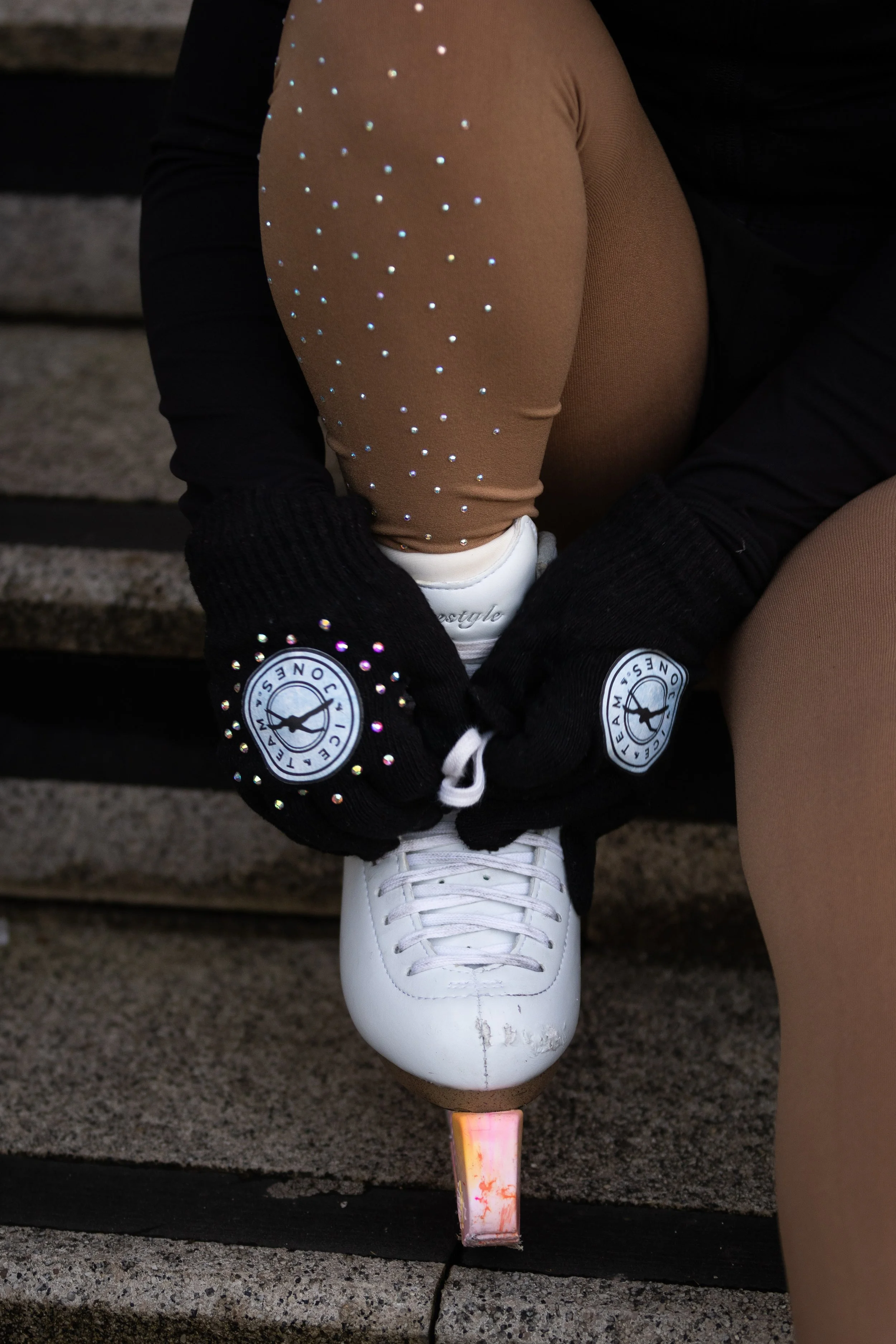 A person tying the skate lace of a white figure skate, wearing tan tights with rhinestones, black gloves with a logo, and black clothing, sitting on concrete stairs.