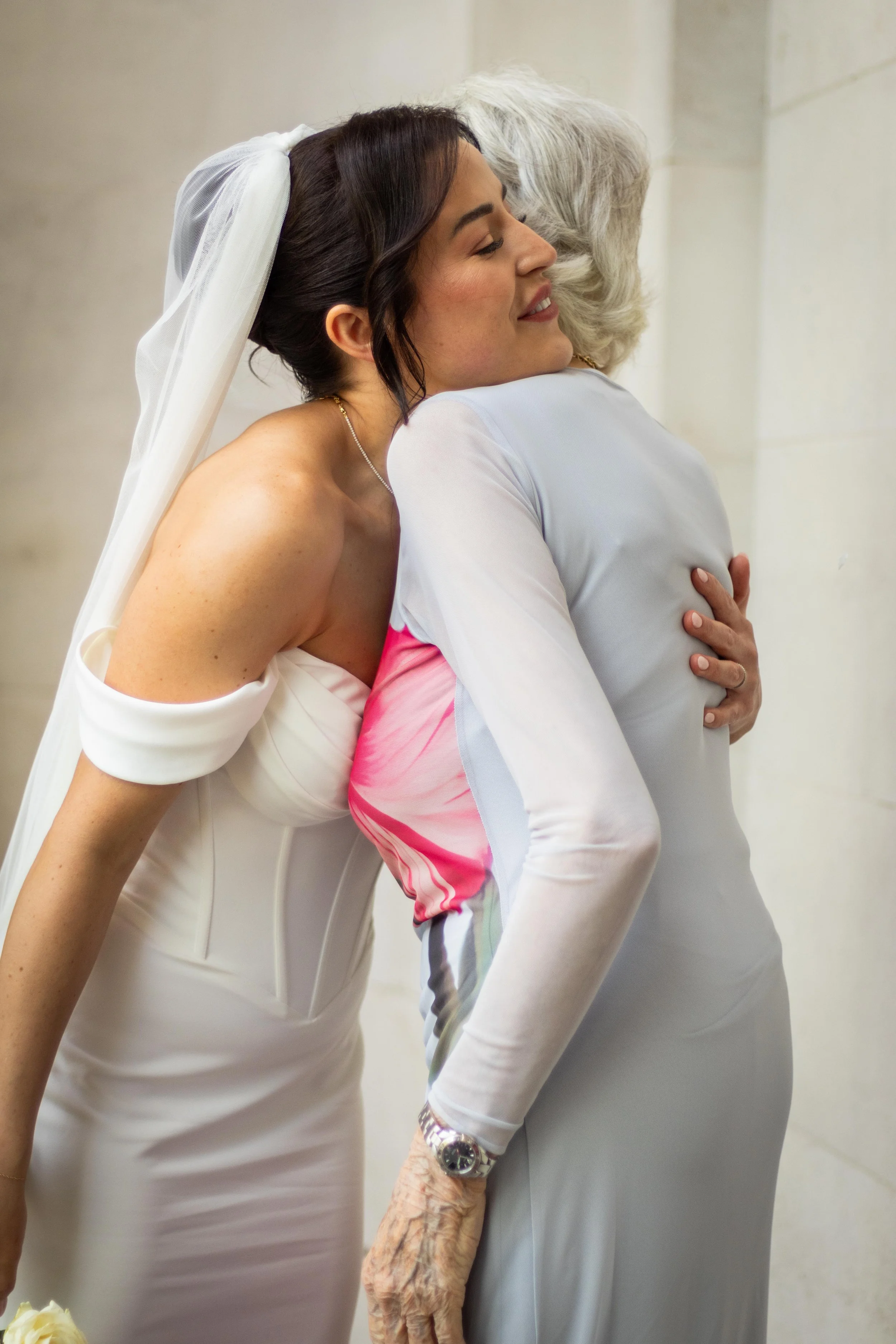 A young woman in a white wedding dress warmly hugs an older woman wearing a light gray dress in an indoor setting with beige walls.