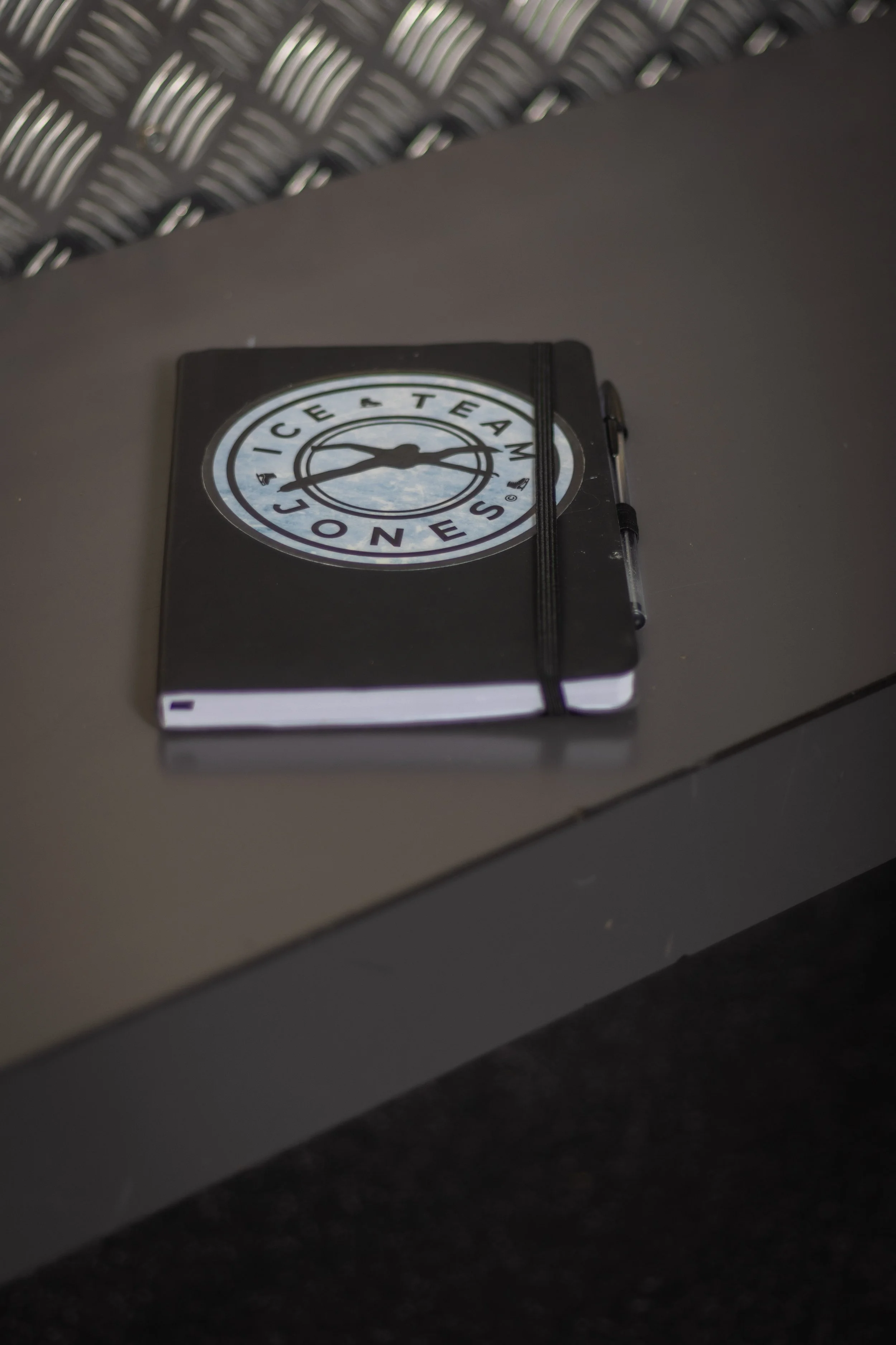 A closed black notebook with the Ice Team sneakers logo on the cover, secured with an elastic band, placed on a dark gray desk with a pen clipped on its side.