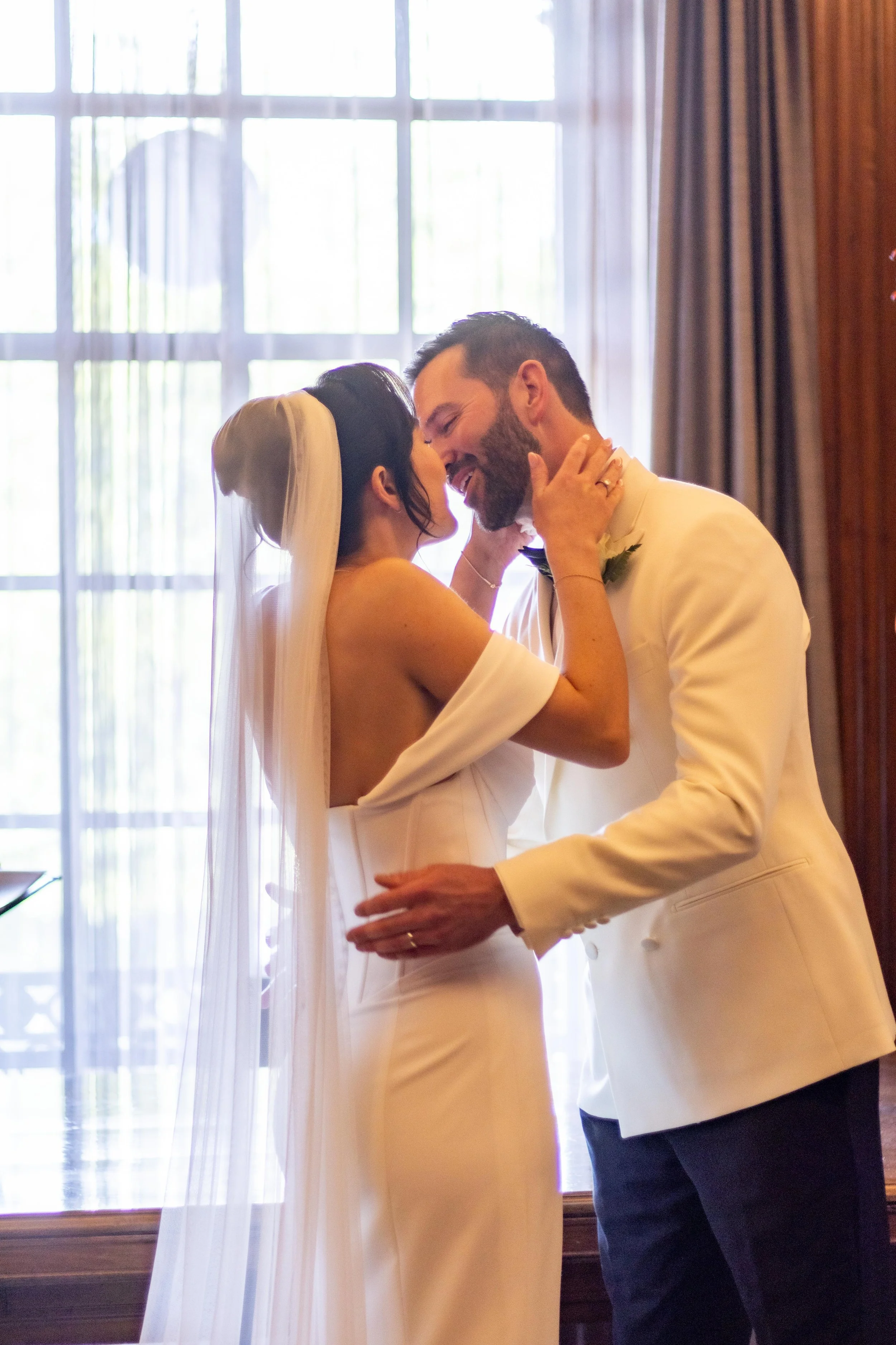 A bride and groom sharing an intimate moment during their wedding ceremony, standing close with faces touching, indoors near a large window with curtains.