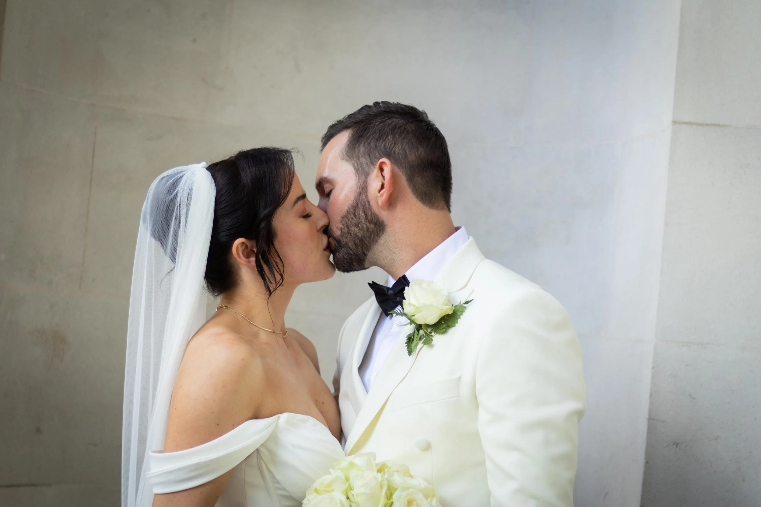 Bride and groom sharing a kiss at their wedding, with the bride wearing a white off-the-shoulder wedding dress and veil, and the groom in a white tuxedo with a black bow tie and a white rose boutonniere, against a neutral wall background.