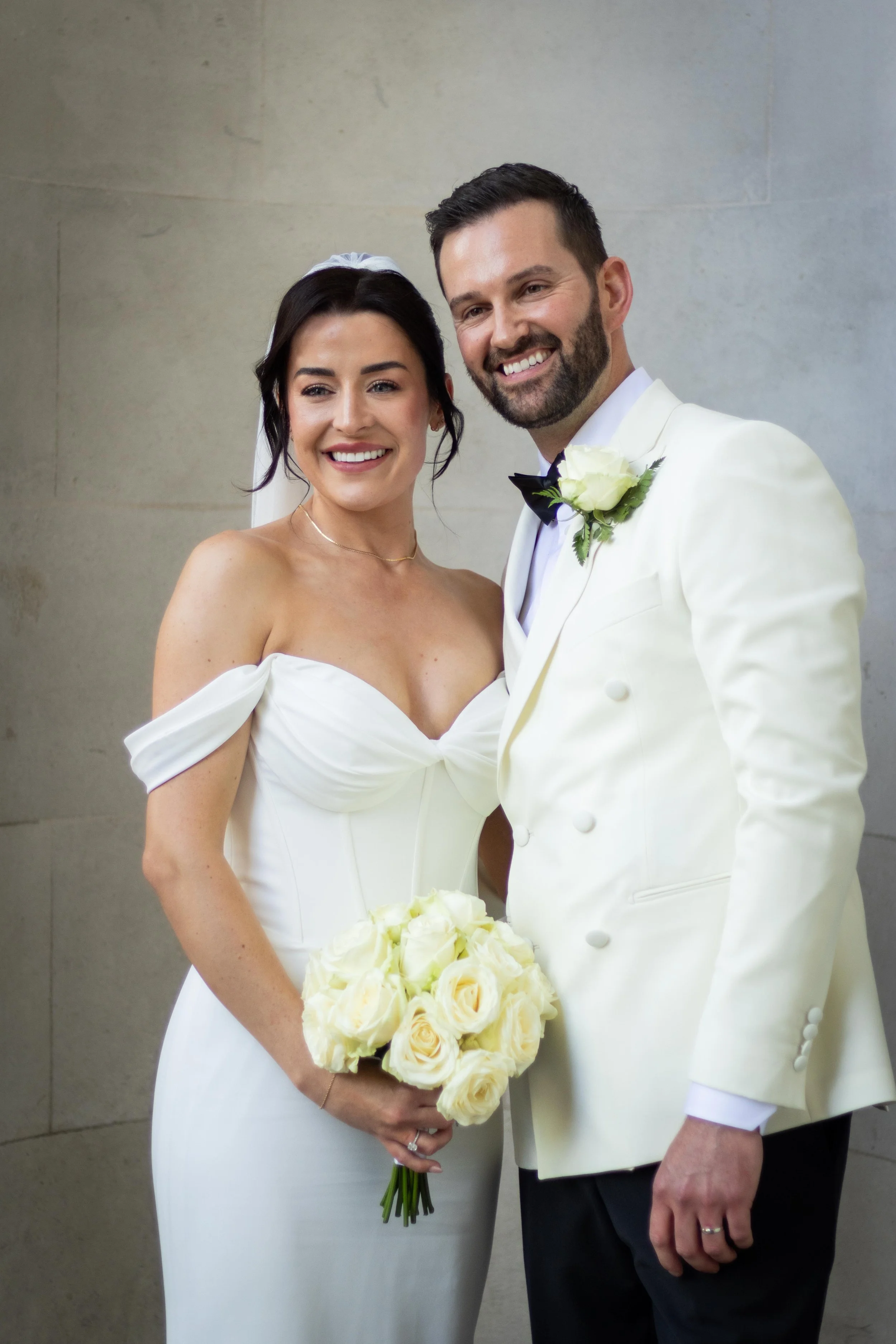 Bride and groom smiling, standing together at their wedding, with the bride holding a bouquet of white roses.