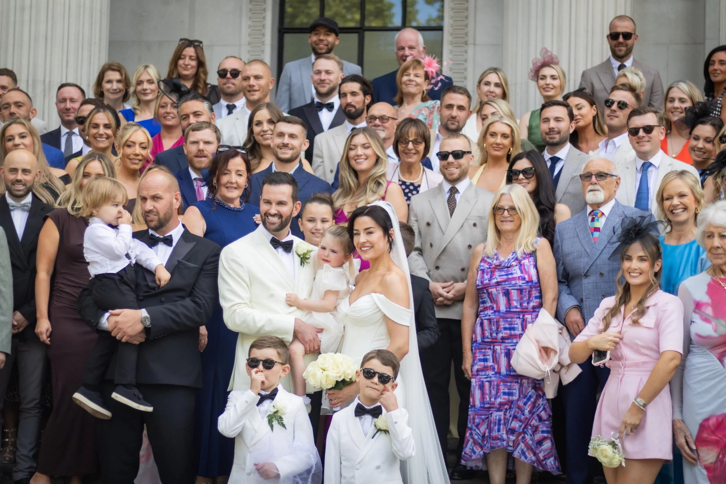 A large group of people gathered for a wedding photo on the steps of a building, with the bride and groom in the center surrounded by family and friends.