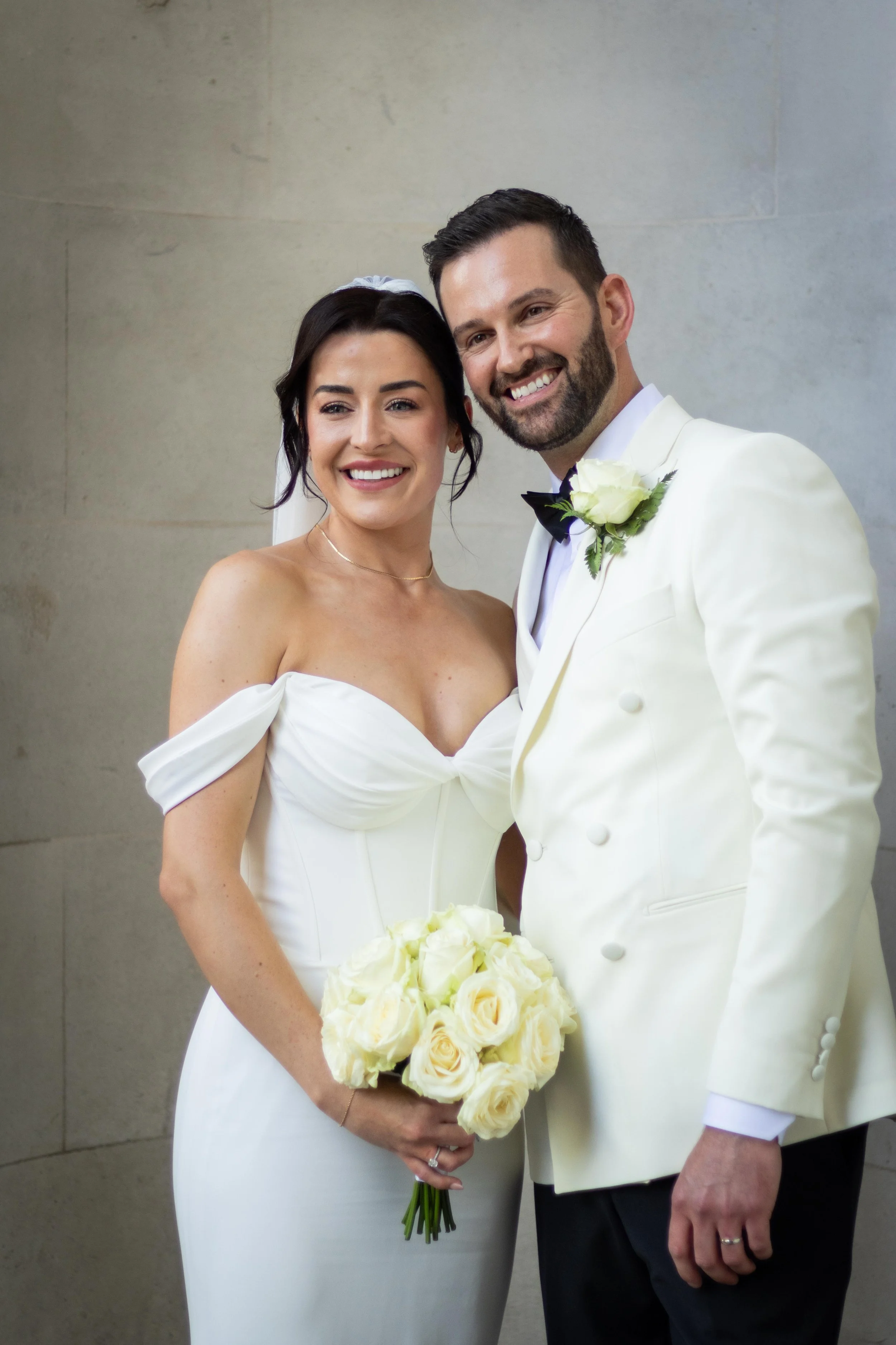 A bride and groom smiling, standing together in wedding attire, with the bride holding a bouquet of white roses.