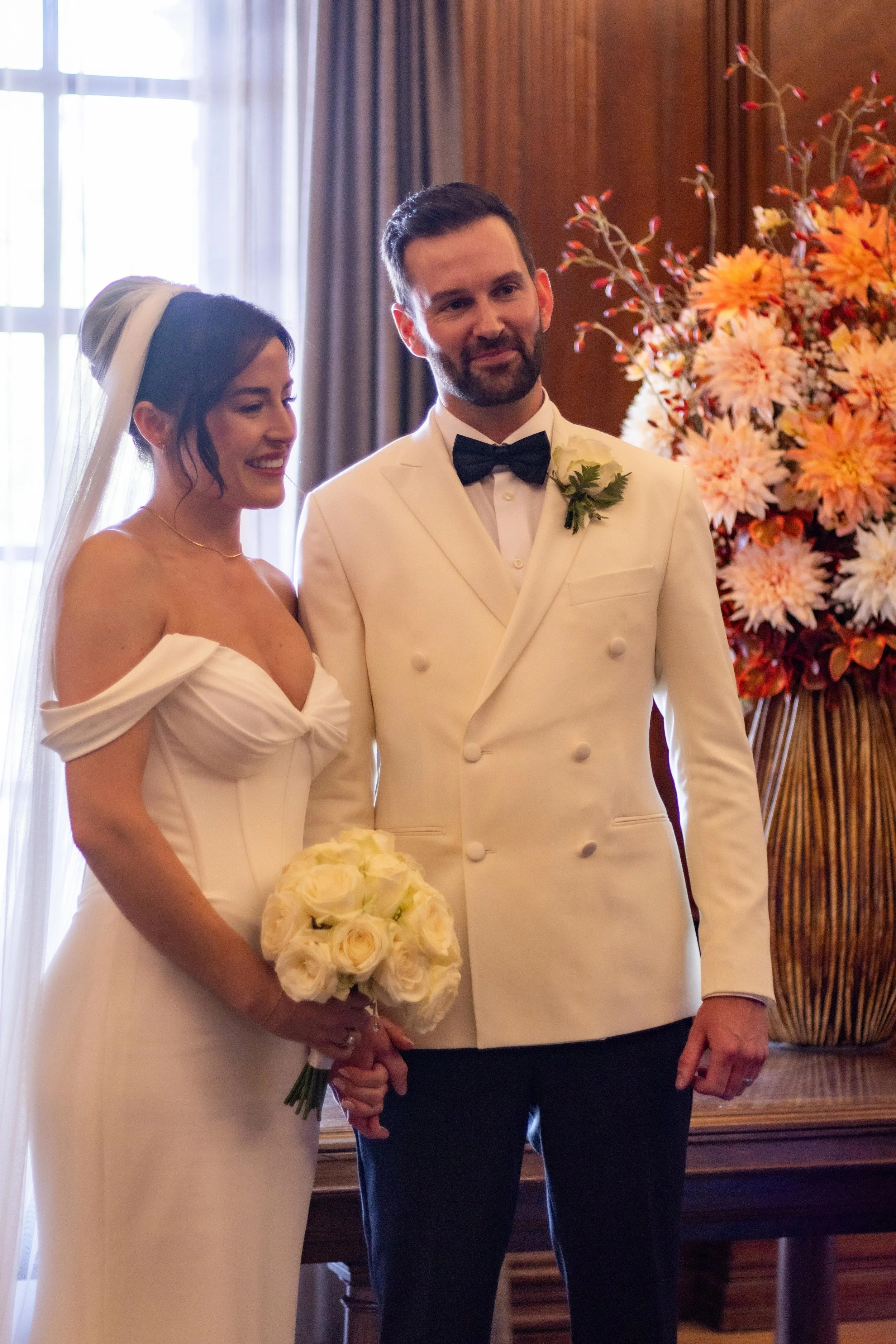 A bride and groom standing together during their wedding ceremony, with the bride holding a bouquet of white roses and the groom in a tuxedo with a bow tie, inside a decorated room with a large floral arrangement in the background.