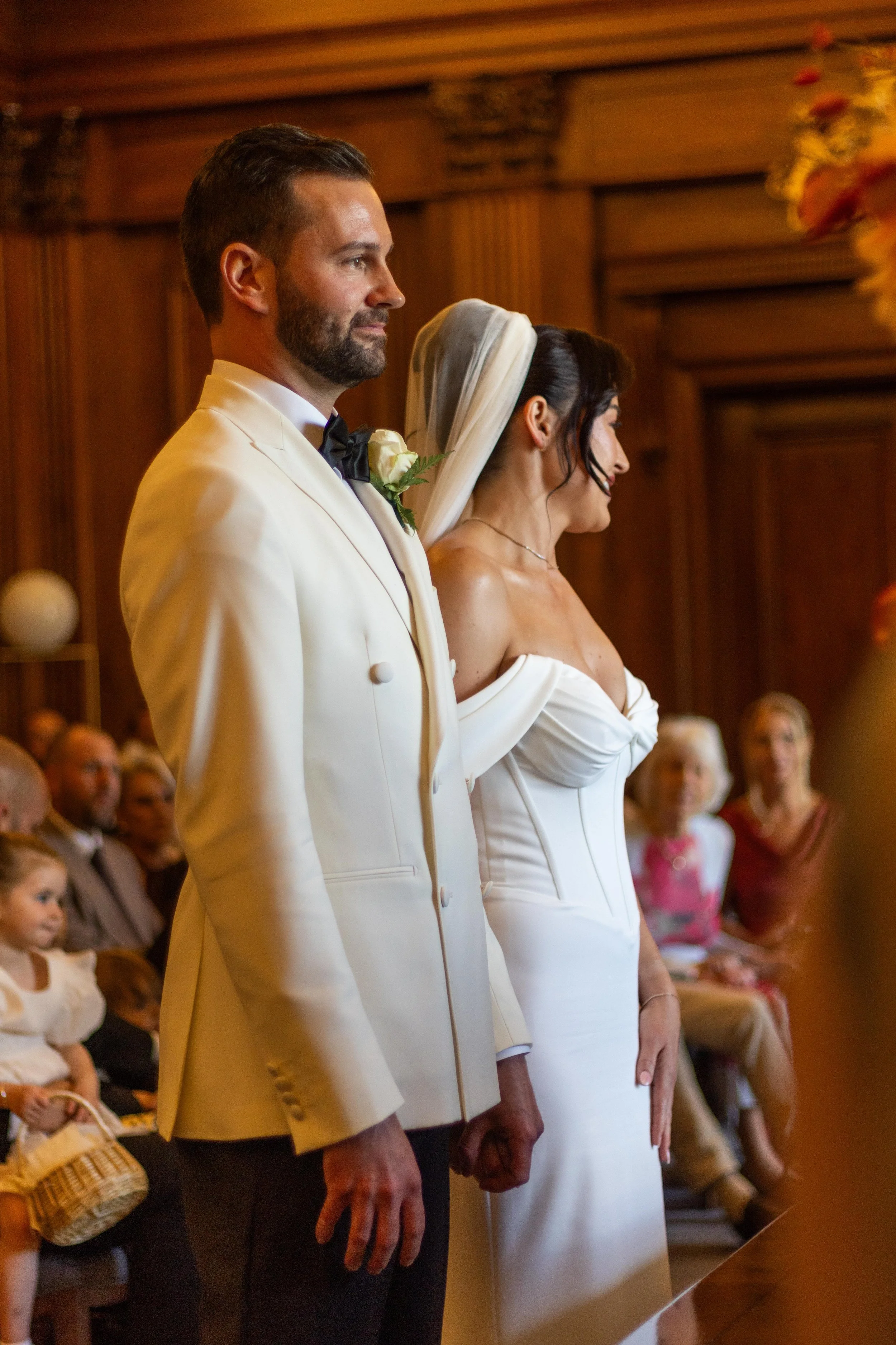 A bride and groom standing during a wedding ceremony, all dressed in white, with guests seated in the background.
