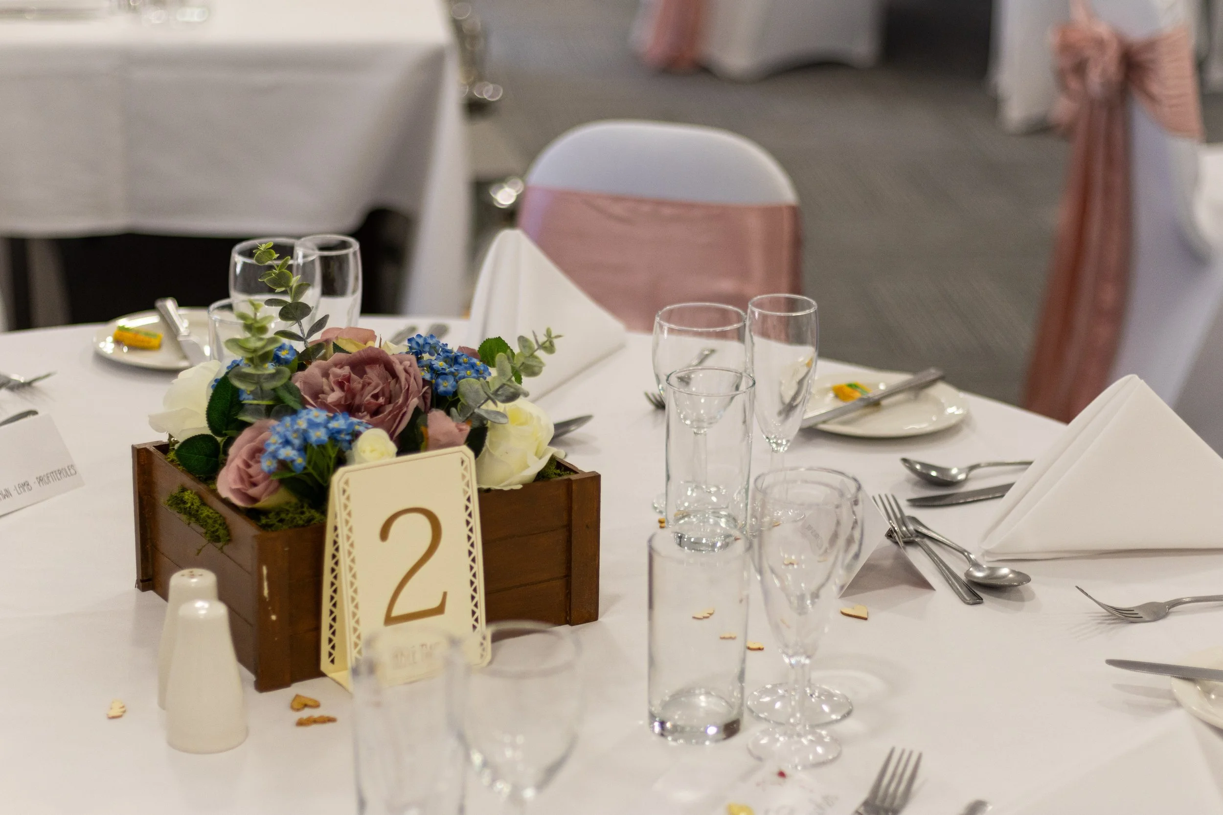 Round banquet table decorated with a floral centerpiece containing pink roses, white roses, blue flowers, and greenery. The table has white tablecloths, folded white napkins, various glassware, silverware, a table number card with the number 2, and s