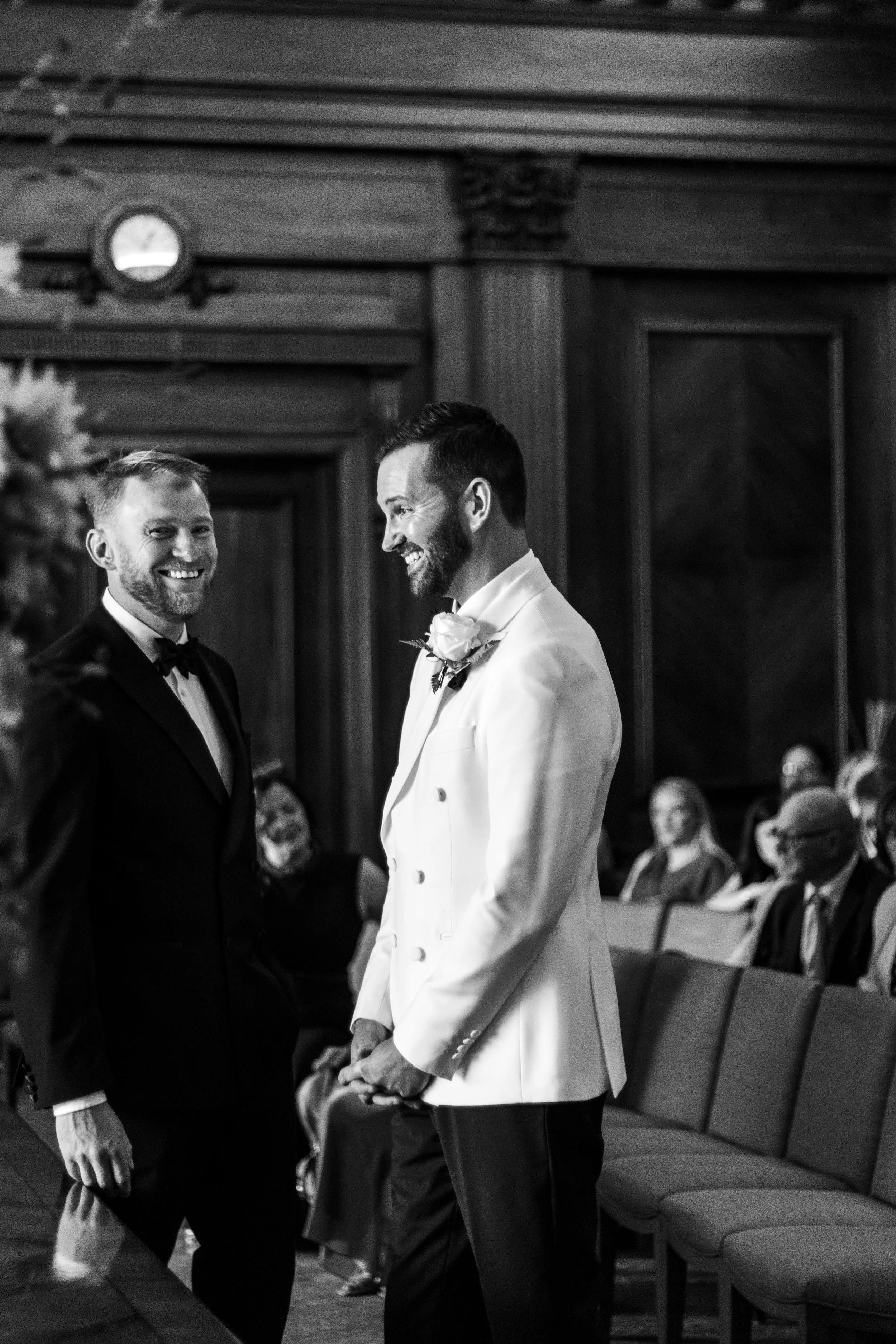A black and white photo of two men at a wedding ceremony, one in a tuxedo and the other in a white suit, smiling at each other. Guests are seated in the background.