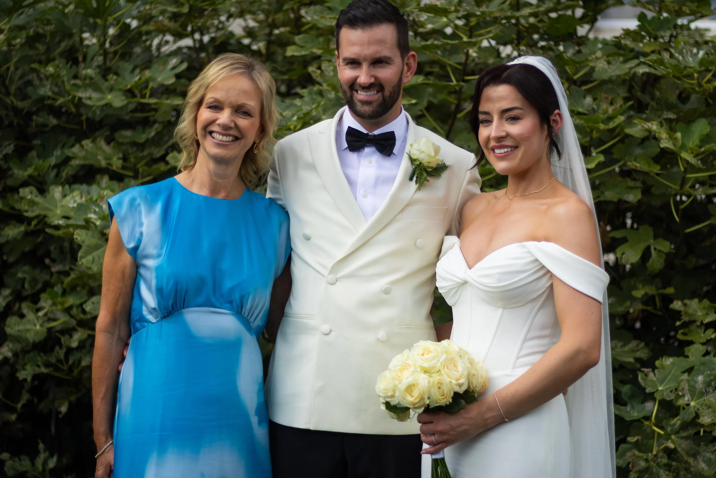 A bride and groom pose with a woman outdoors, with greenery in the background. The bride wears a white off-shoulder gown with a veil, holding a bouquet of white roses. The groom wears a white tuxedo with a black bow tie. The woman wears a bright blue