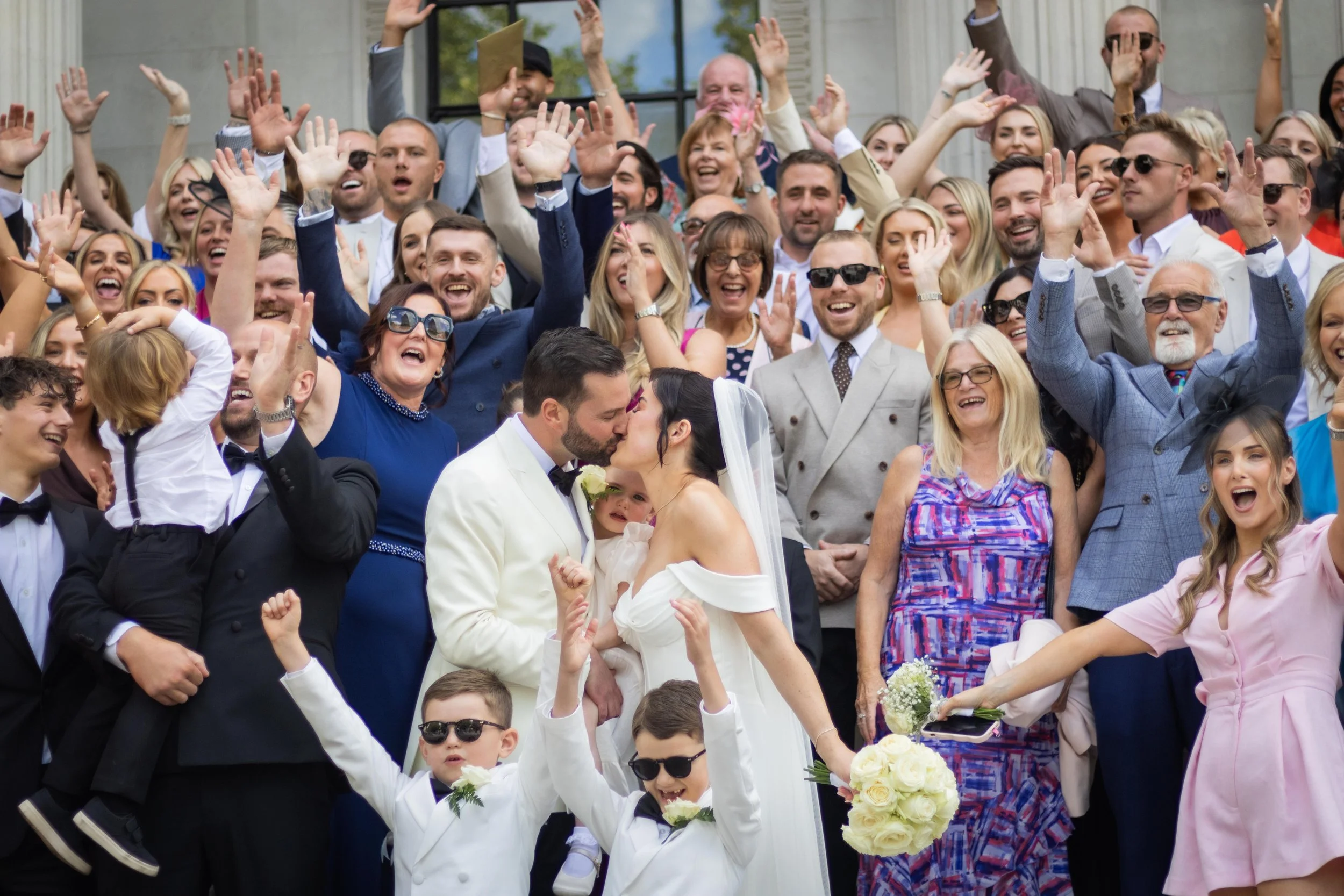 A large group of people celebrating at a wedding, with the bride and groom sharing a kiss in the front, surrounded by friends and family cheering and raising their hands.