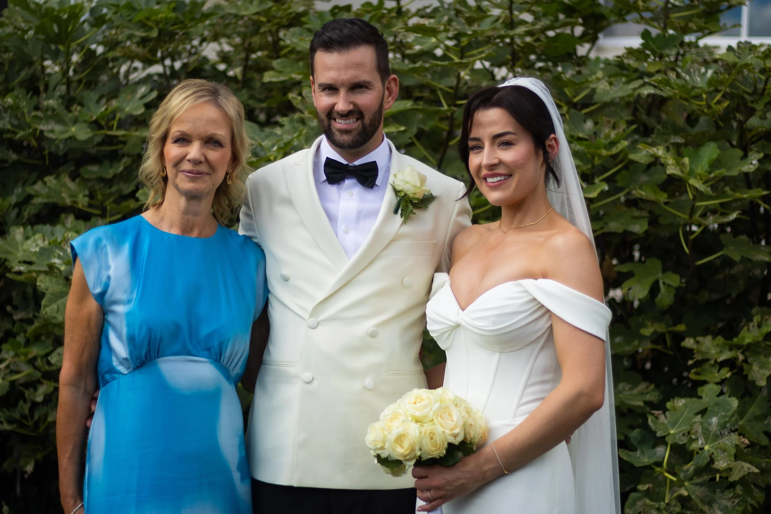 A bride and groom with a woman, smiling outdoors in front of greenery, at a wedding.