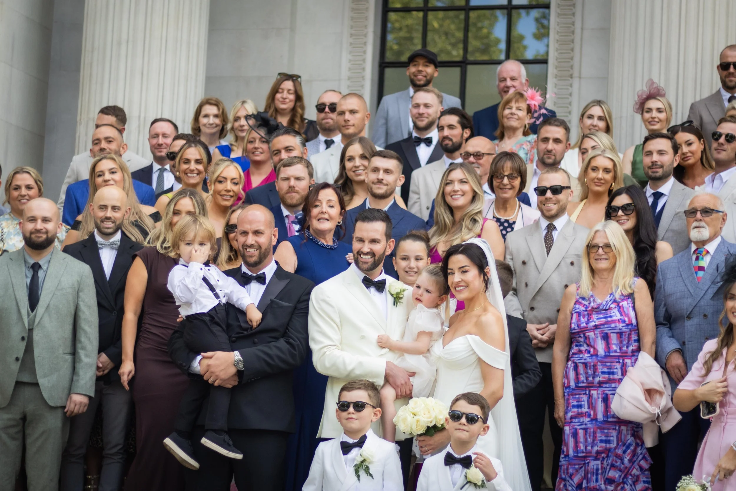 A large group of people in formal attire gathered for a wedding photo on the steps of a building with columns, smiling and posing for the camera.