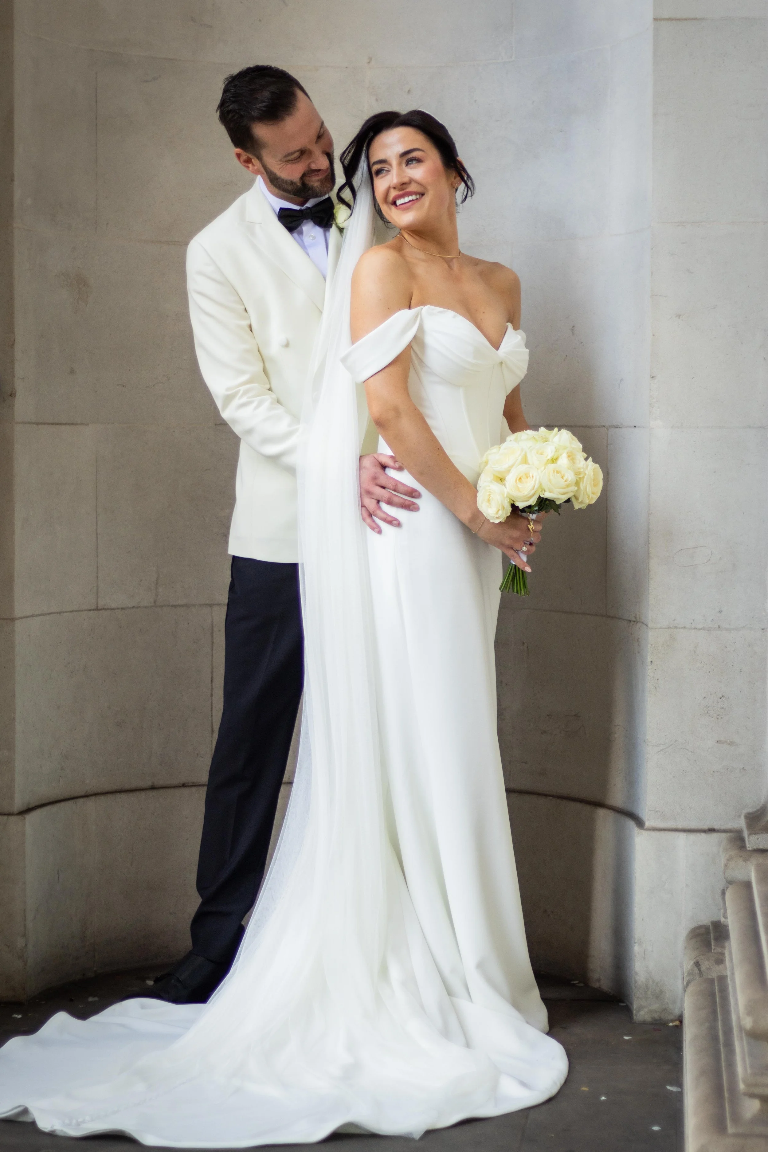 A newly married couple, the groom in a white tuxedo jacket and black bow tie, and the bride in a white off-shoulder wedding gown, standing close together with the groom embracing the bride from behind, holding a bouquet of white roses, against a plai