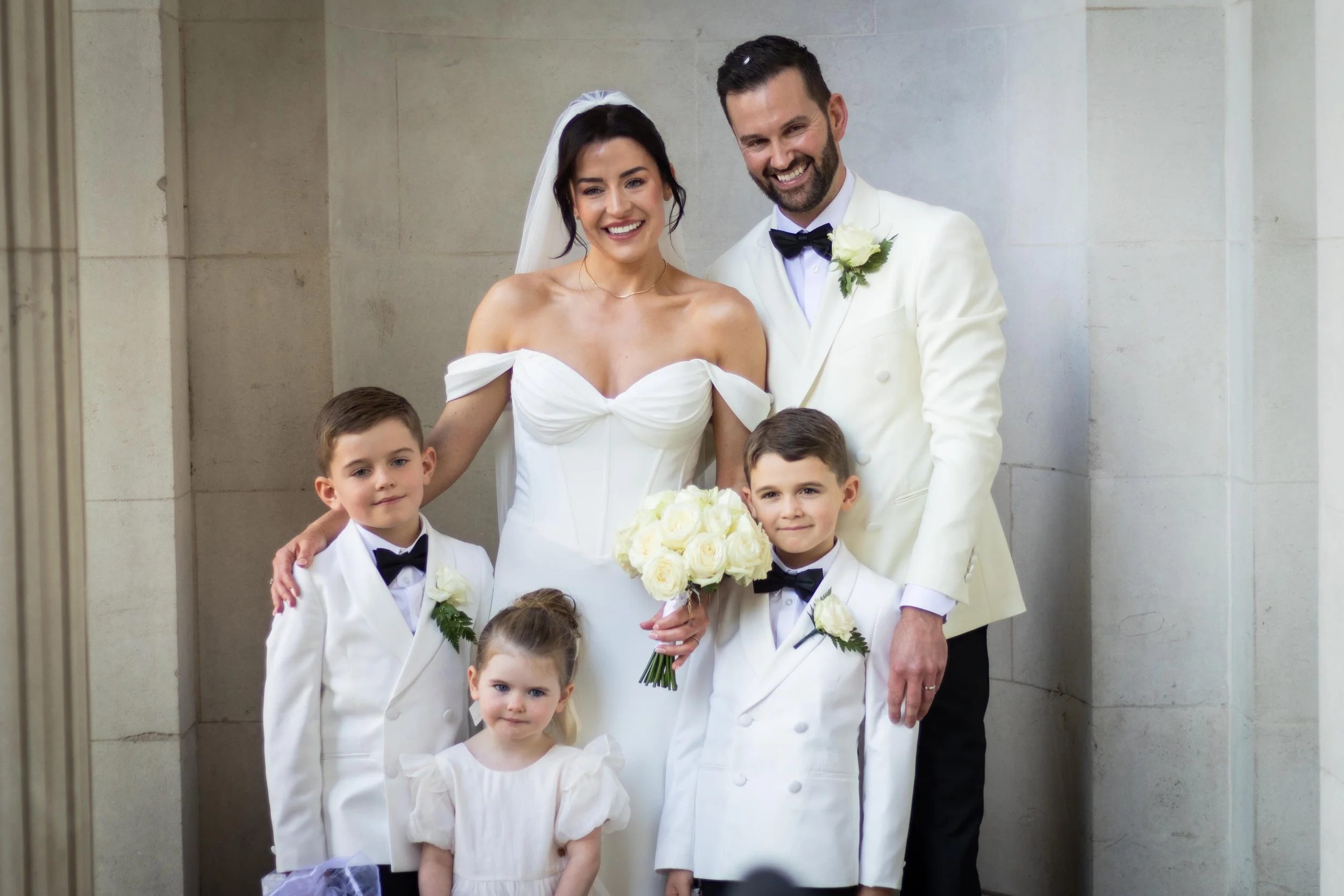 A wedding party with a bride in a white dress holding a bouquet of white roses, a groom in a white tuxedo with a black bow tie, and four children dressed in white with black bow ties, standing against a stone wall.