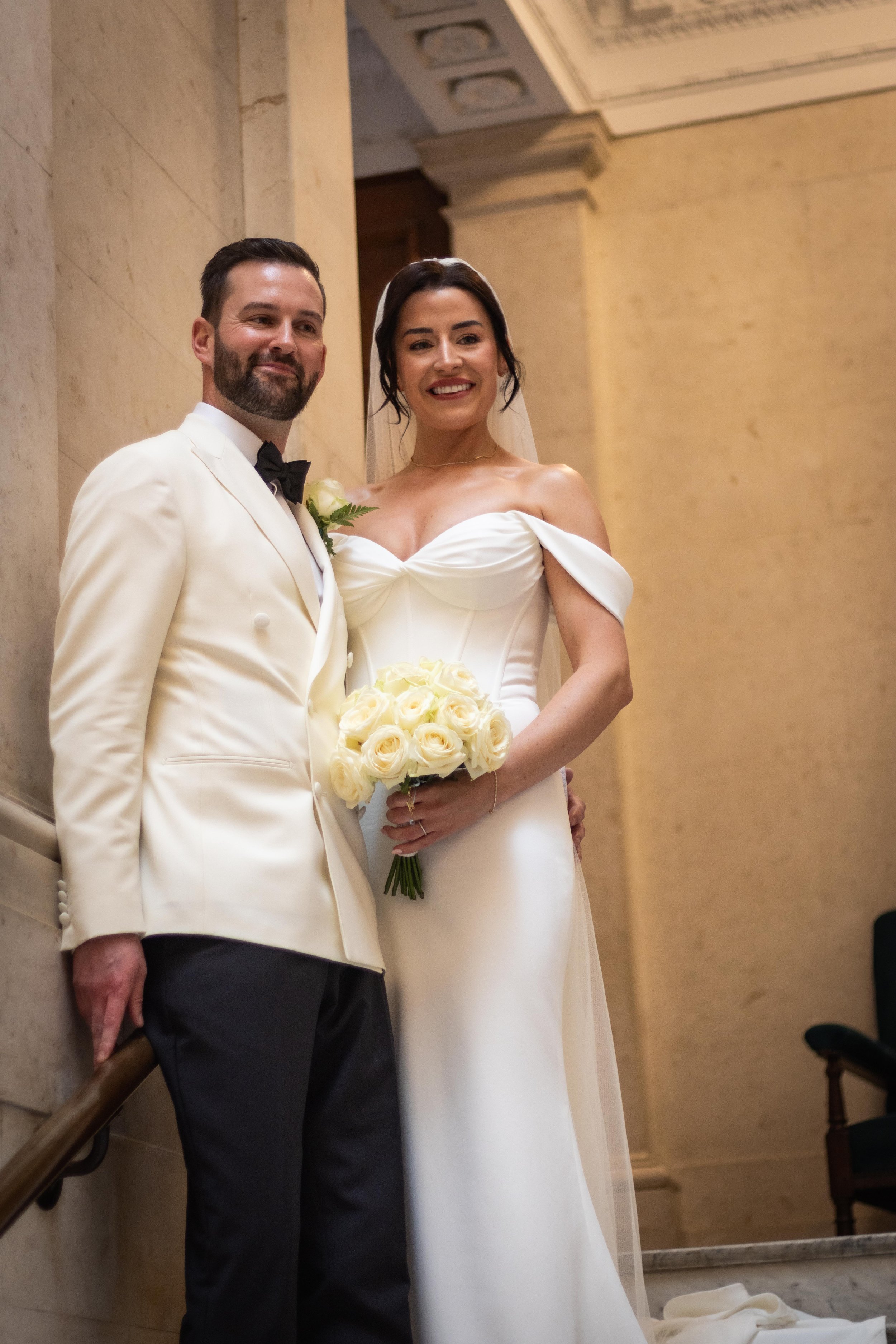 A newlywed couple in wedding attire posing indoors, with the bride holding a bouquet of white roses.