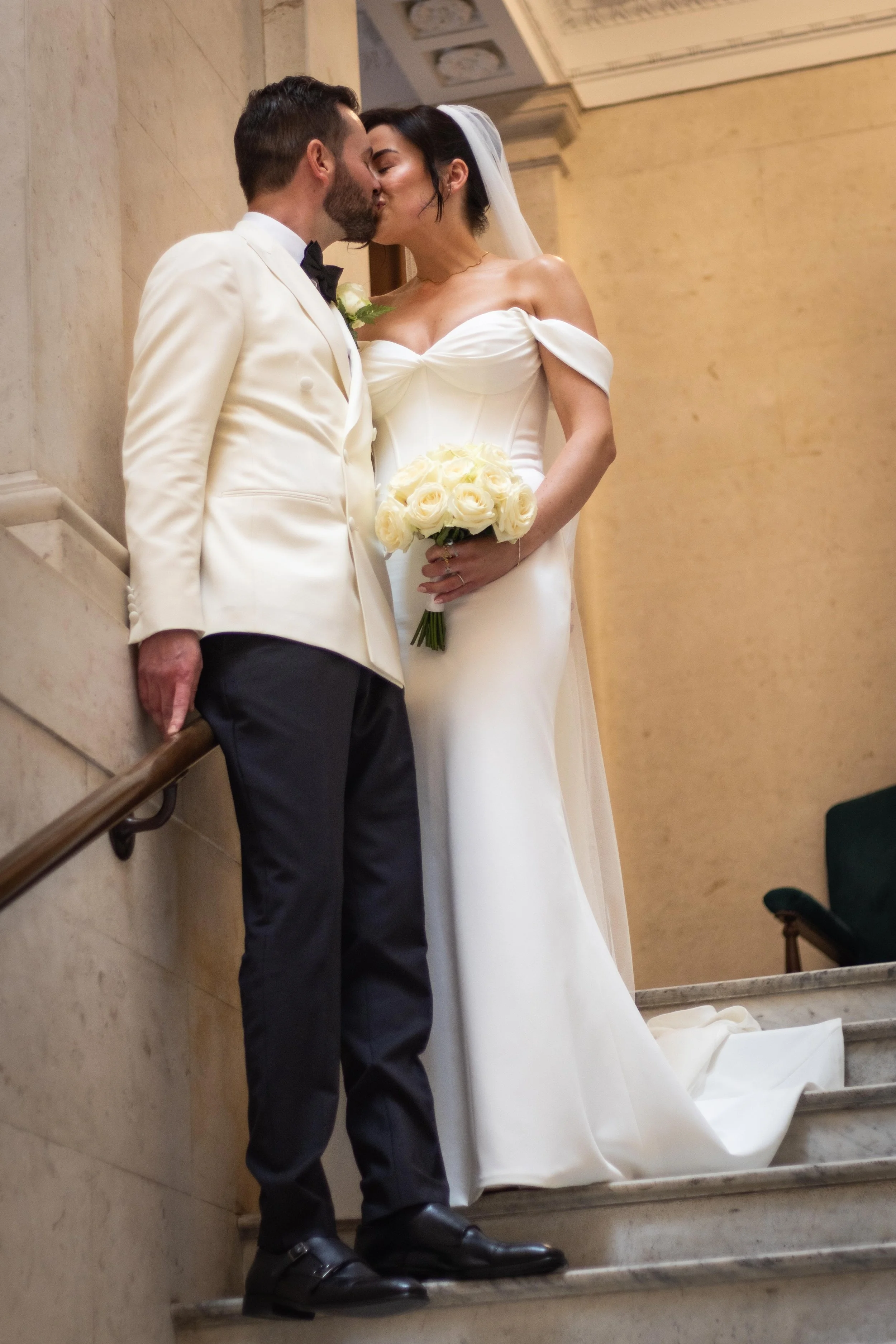 A bride and groom share a kiss on the stairs during their wedding, with the bride holding a bouquet of white roses.