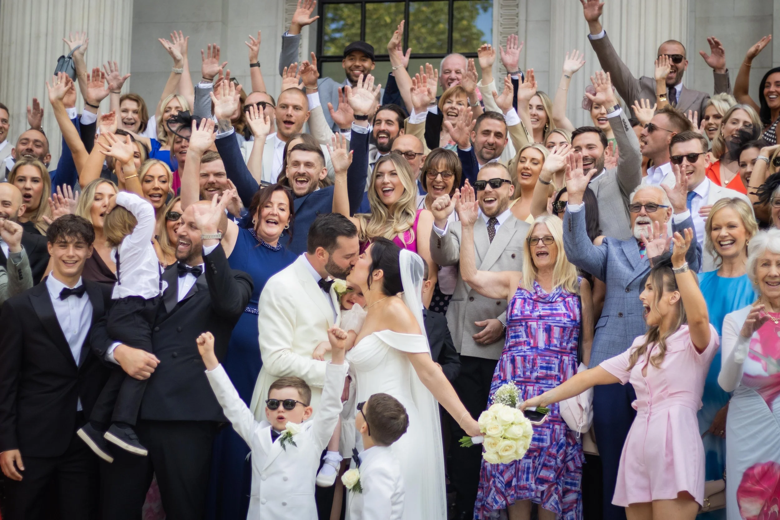 A large group of people at a wedding celebration, including the bride and groom kissing, surrounded by smiling and cheering guests.